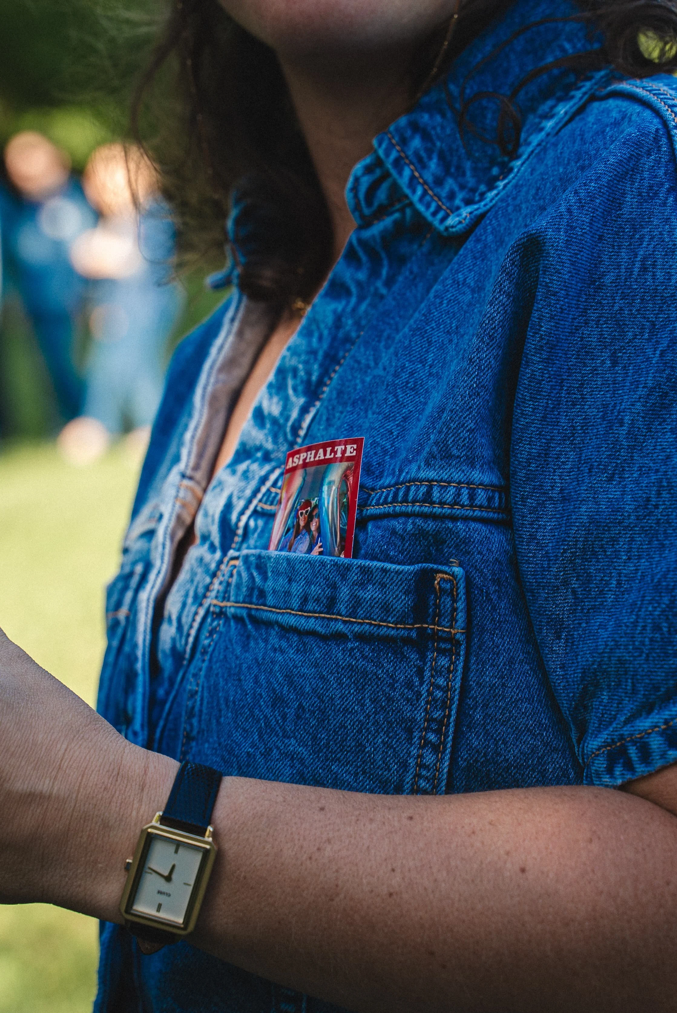 Une personne porte une veste en jean et un bracelet noir, avec une montre à cadran carré et bracelet en cuir noir. La personne tient dans la main une carte postale en plastique avec une photo de personnes et le mot 'Asphalte'.