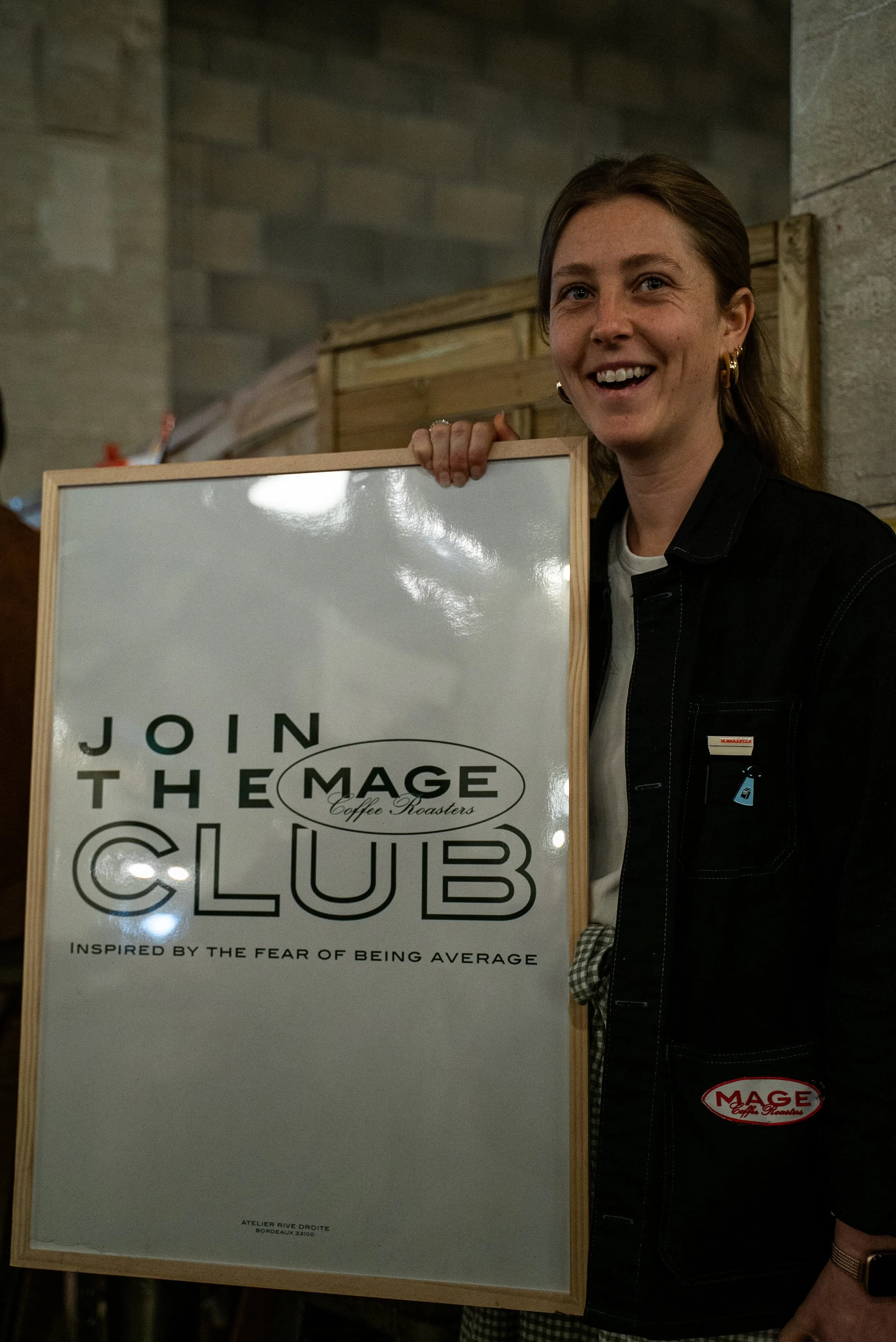 Une femme souriante tenant un tableau blanc avec un panneau publicitaire annonçant une réunion du club 'Join the Magic', inspirée par la peur d'être ordinaire, située à Bordeaux, France.