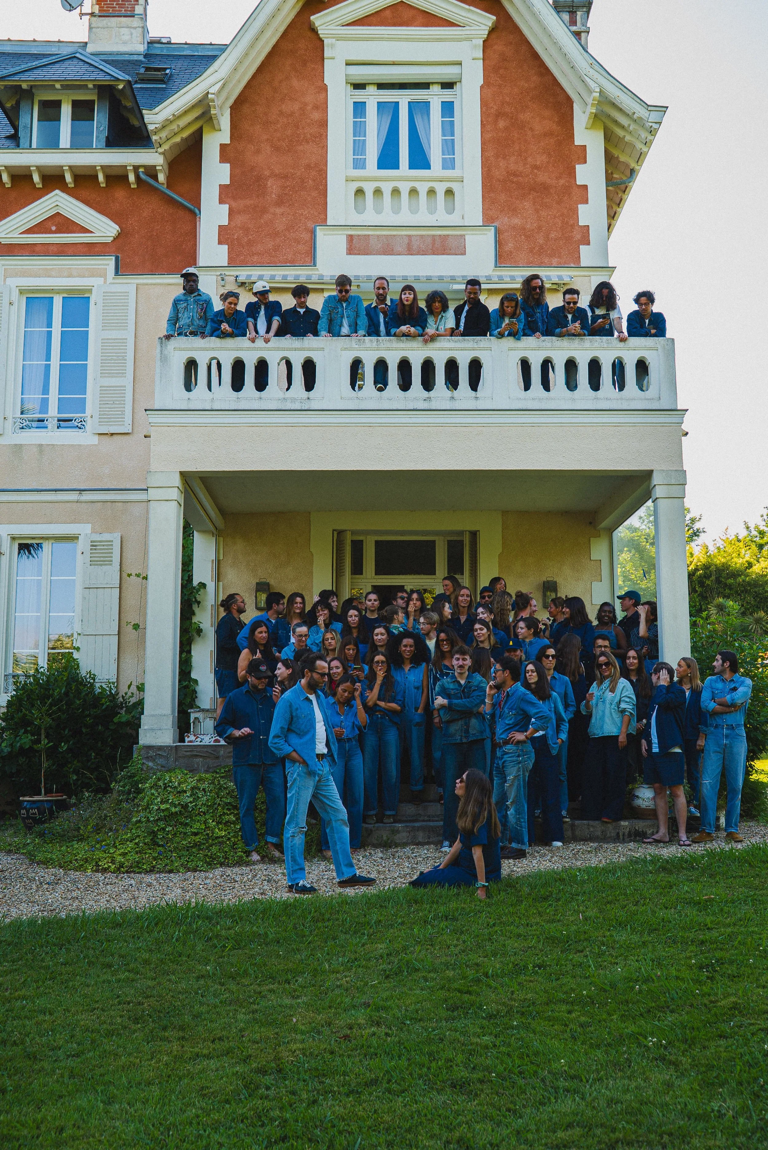 Un groupe de personnes, principalement jeunes adultes, se rassemble sur la terrasse d'une maison en pierre colorée, avec beaucoup de fenêtres et un balcon, par une journée ensoleillée.