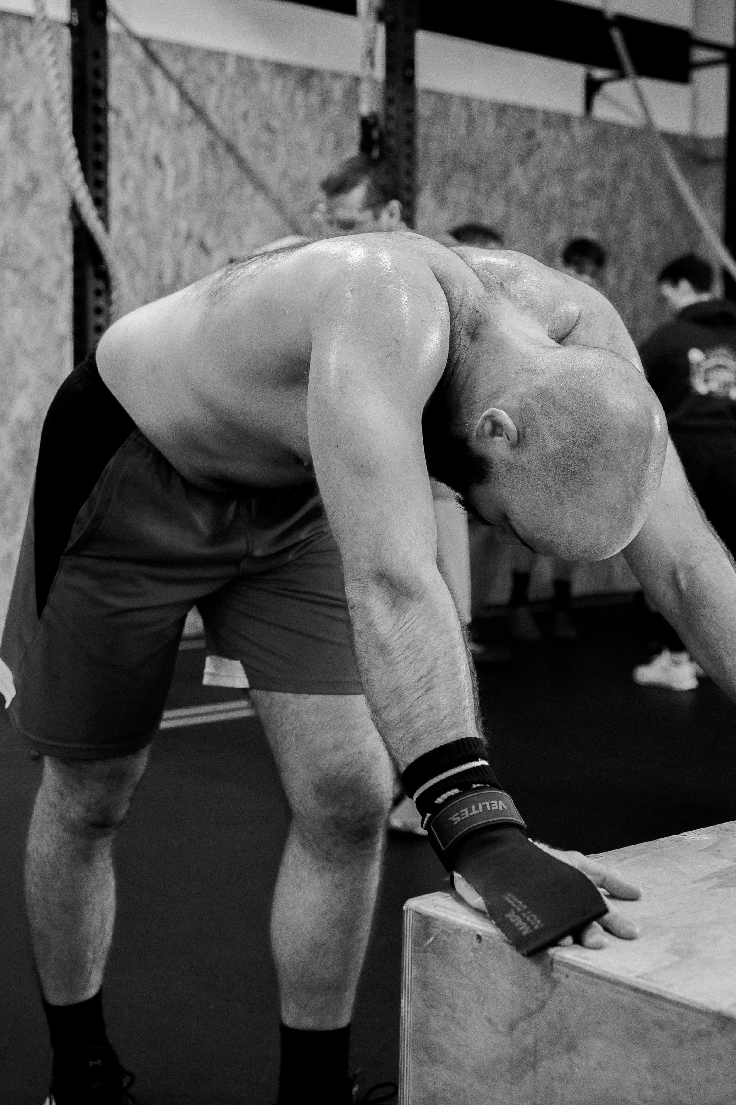 Un homme musclé en short noir, portant des bandages aux poignets, qui s'apprête à soulever un objet lourd en position de squat dans une salle de sport.