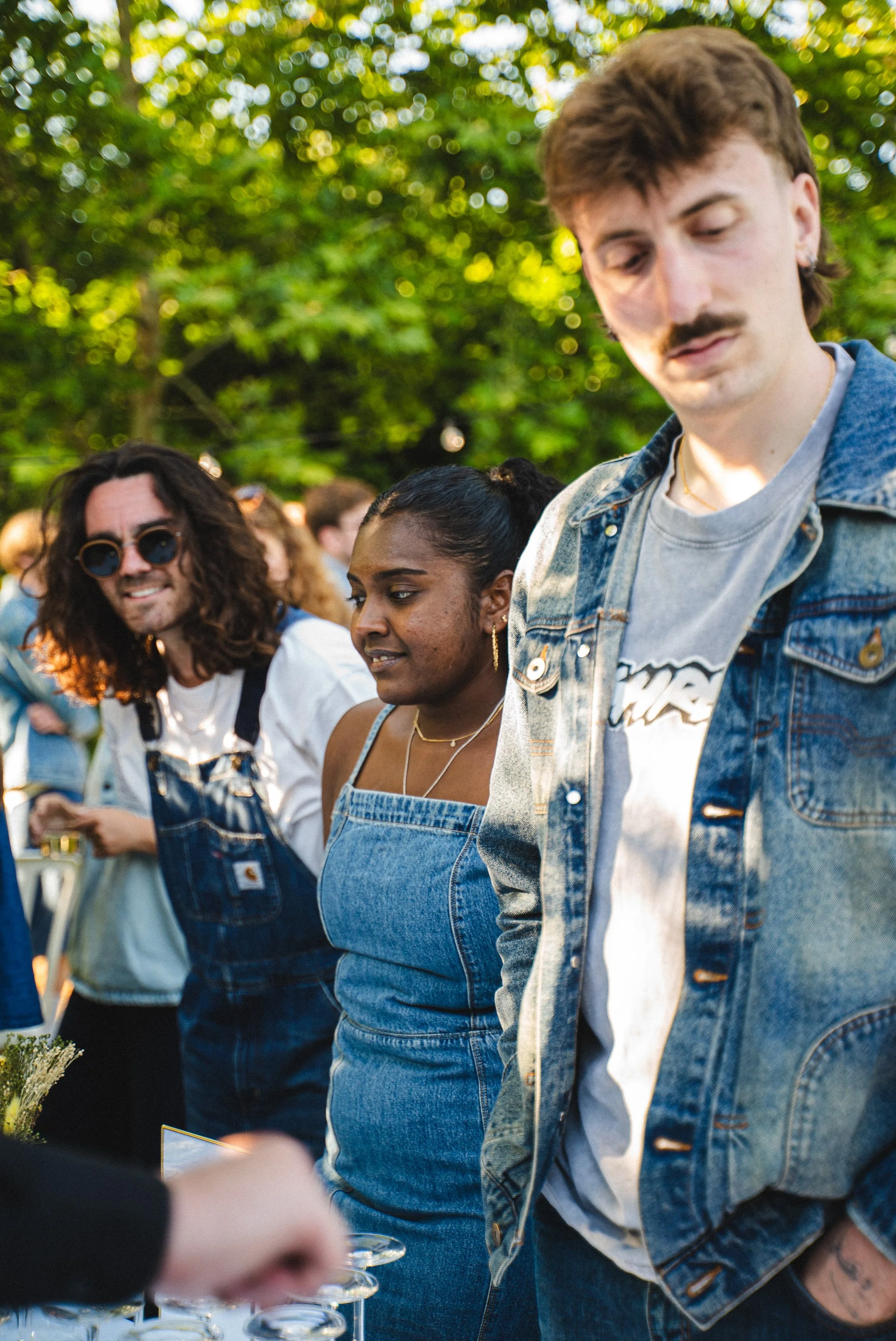 Groupe de jeunes adultes lors d'un rassemblement extérieur, certains portant des vêtements en denim, entourés d'arbres et de lumière naturelle.