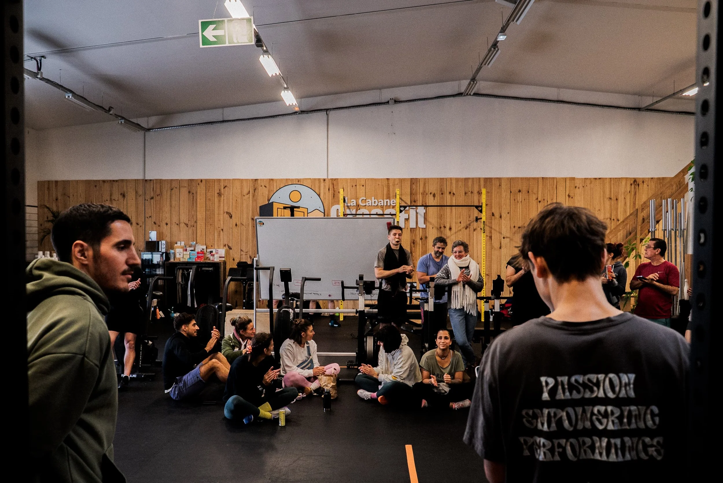 Un groupe de personnes dans une salle de sport, certaines sont assises par terre, d'autres debout, dans une ambiance détendue. La salle a un panneau lumineux vert avec une flèche, un mur en bois derrière avec le logo 'la Cabane CrossFit'.