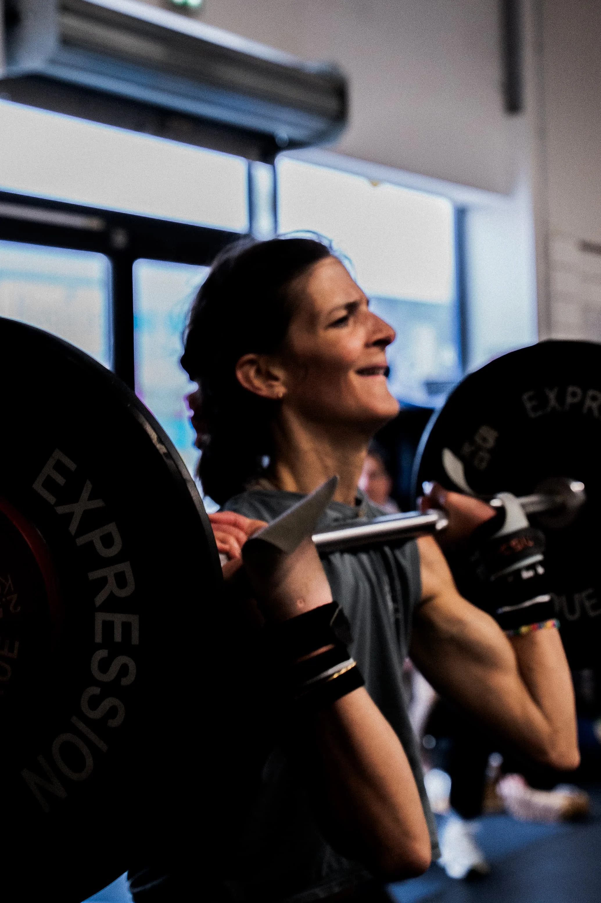 Une femme soulève une barre d'haltérophilie dans une salle de sport, souriant.