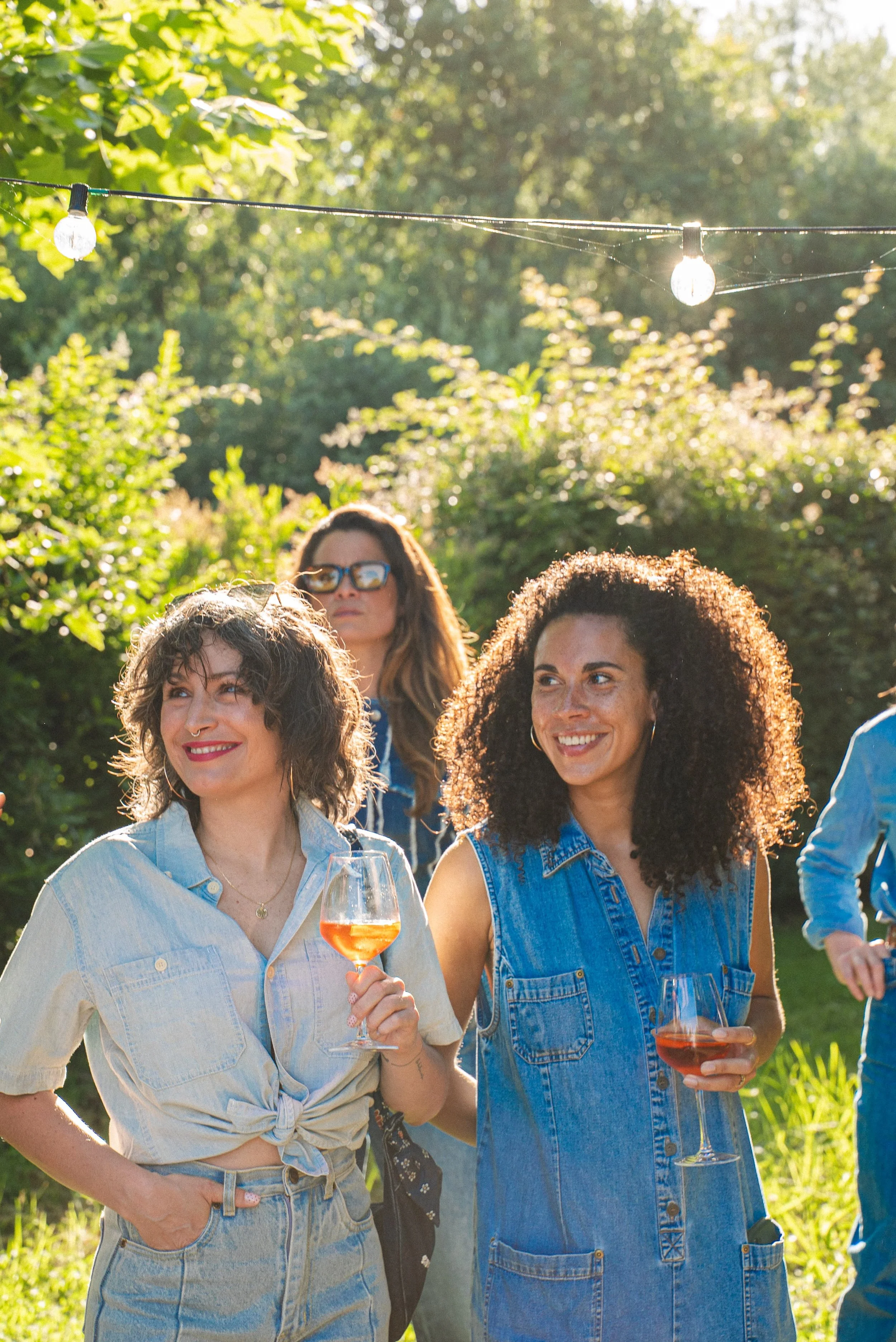 Un groupe de femmes souriantes en plein air, tenant des verres de vin rosé lors d'une dégustation ou d'une fête dans un jardin ensoleillé avec des lumières suspendues.
