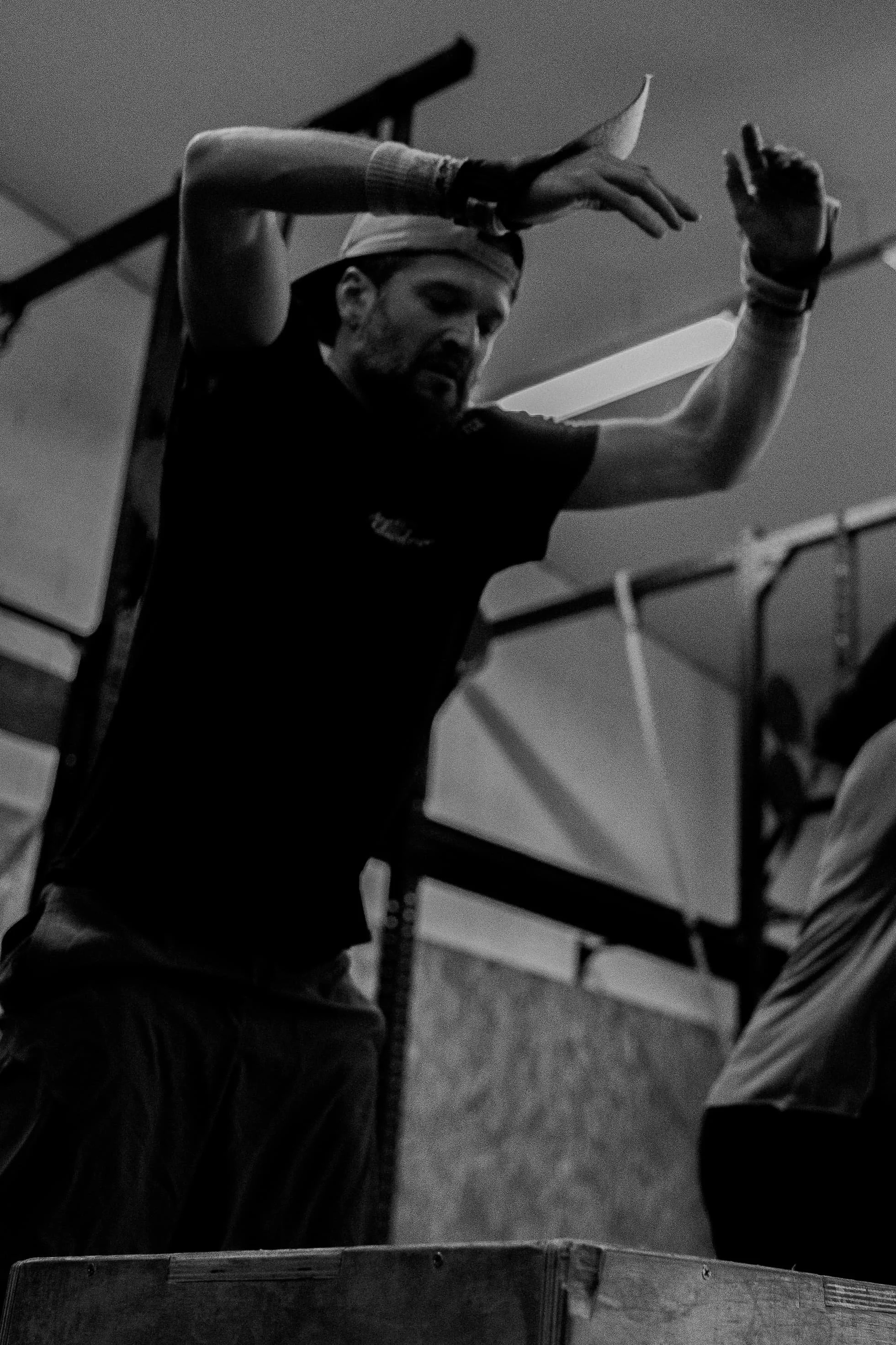 Un homme en t-shirt noir, portant un bandeau blanc et des bracelets noirs, dans une salle avec un plafond bas, en train de faire un geste avec ses mains pendant qu'il se tient derrière une barrière en bois.
