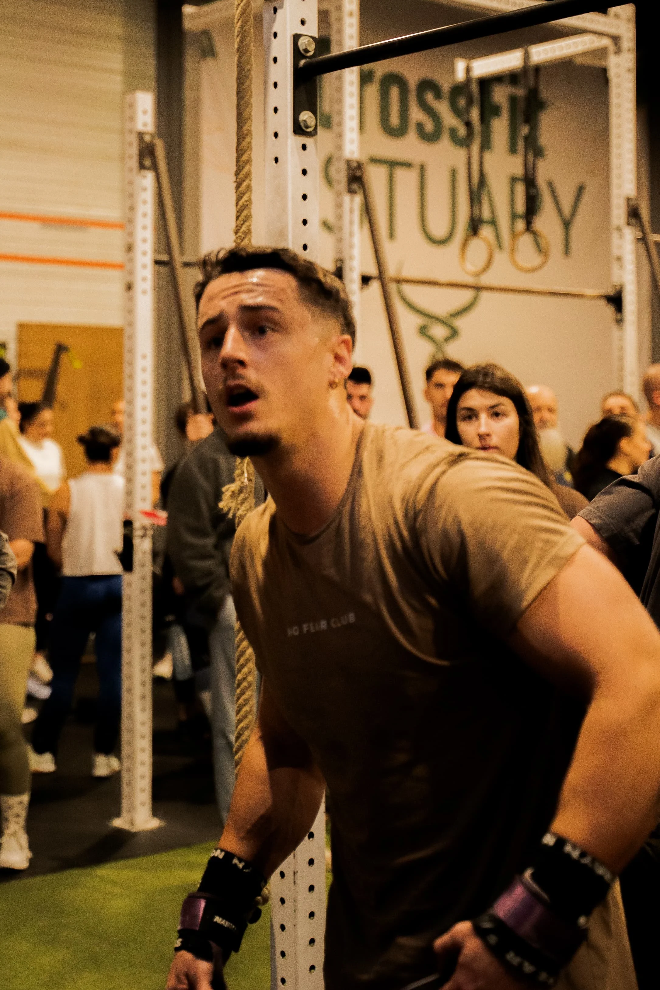 Un homme en train de faire un entraînement de musculation ou de CrossFit dans une salle, entouré d'autres personnes. L'homme porte des vêtements de sport et des poignets de soutien, avec une structure de rameur ou de matériel de gymnastique visible e