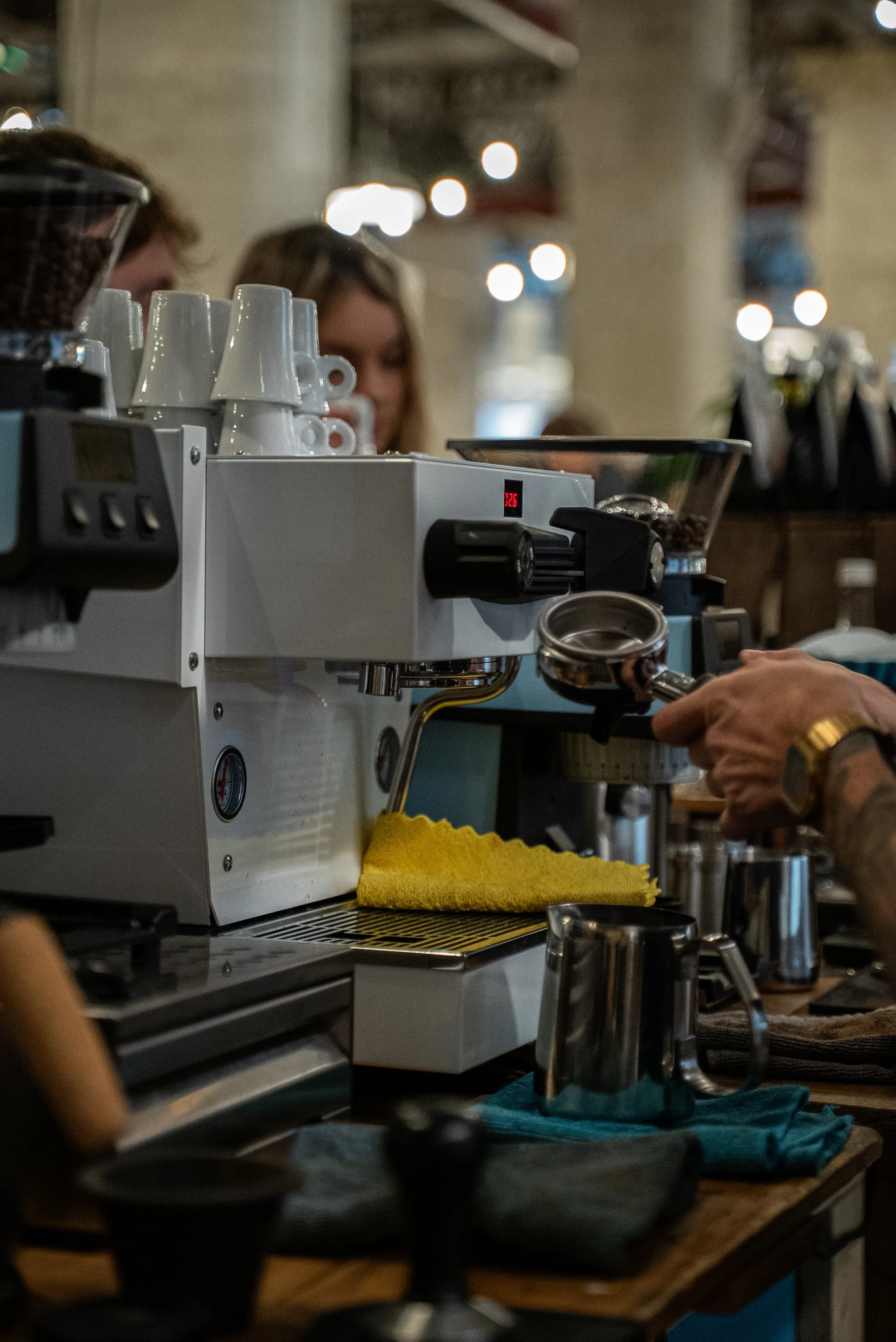 Une machine espresso avec des tasses blanches empilées dessus, un moulin à café, une tasse en métal vide et un chiffon en tissu bleu sur un comptoir en bois dans un café.