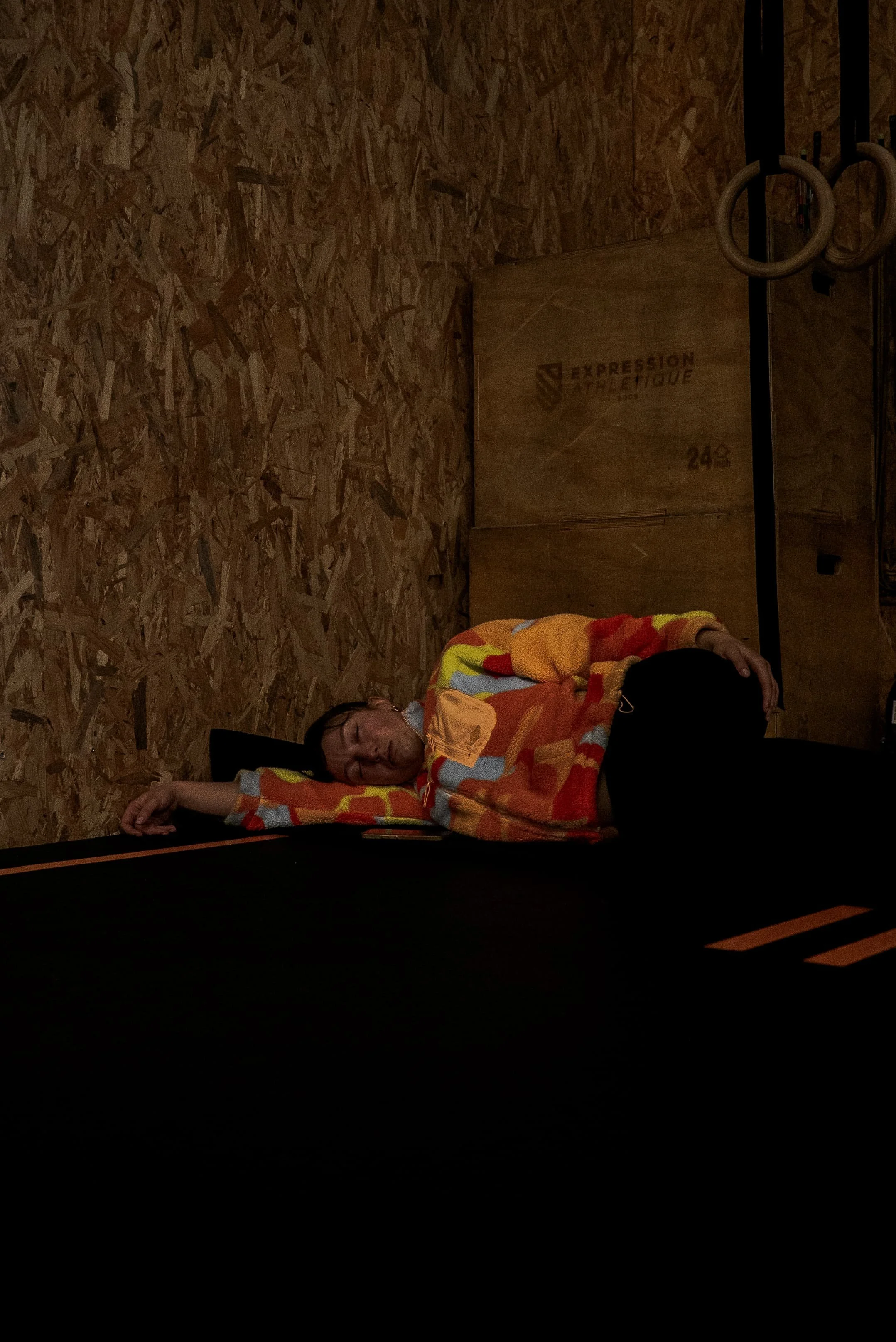 Une personne repose sur un banc dans une salle de sport avec murs en panneaux de bois, portant un sweat à motifs orange, rouge, jaune. Il y a des anneaux de gymnastique suspendus au mur.