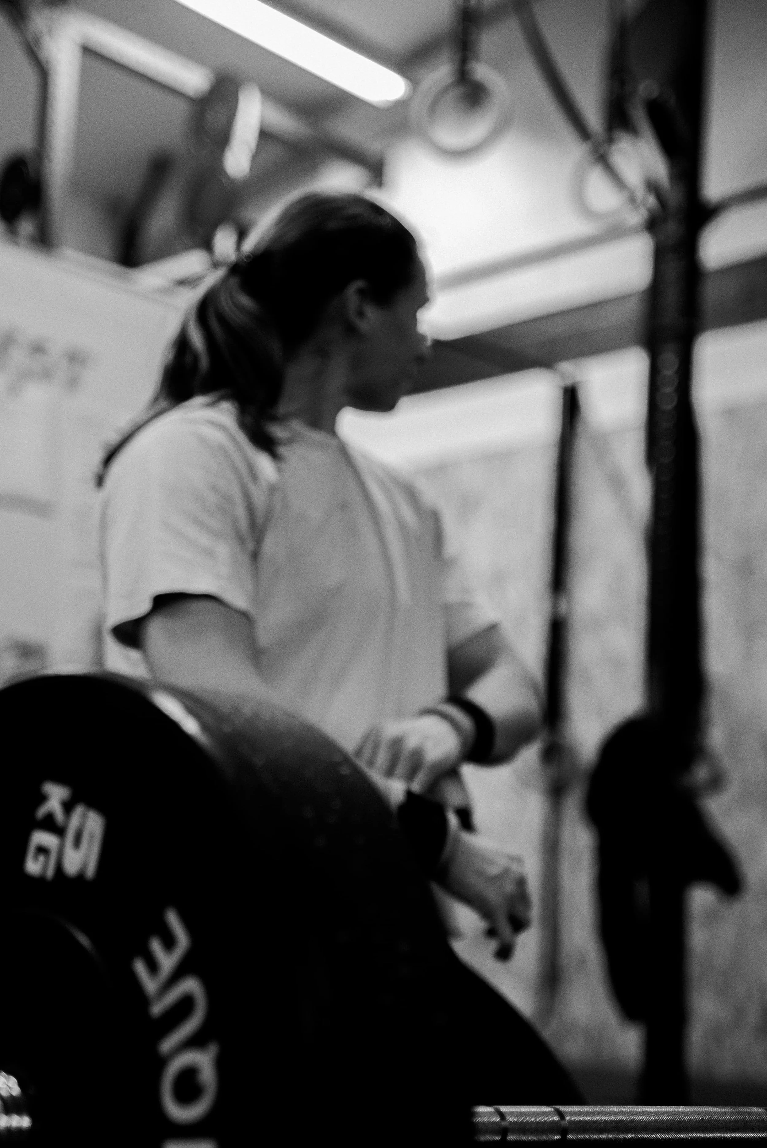 Une femme portant un t-shirt blanc, vue de profil, se tient à côté d'un rack de haltères dans une salle de sport ou une zone d'entraînement.
