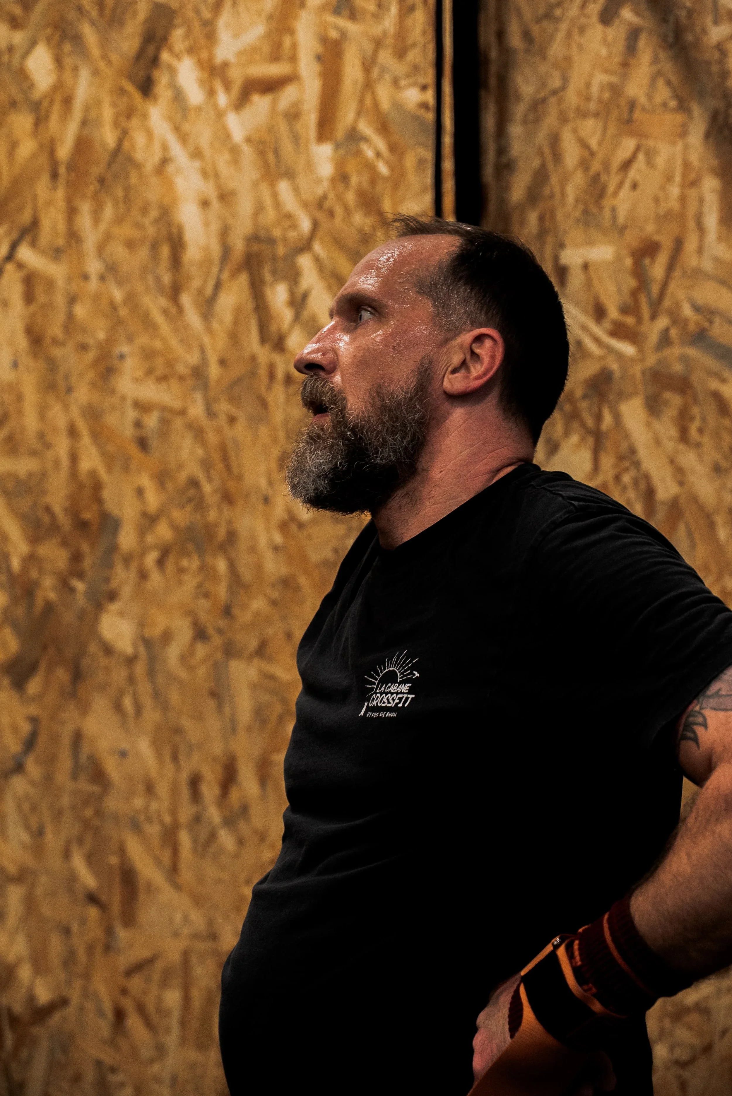 Profil d'un homme avec une barbe, portant un t-shirt noir avec un logo, contre un mur en bois.