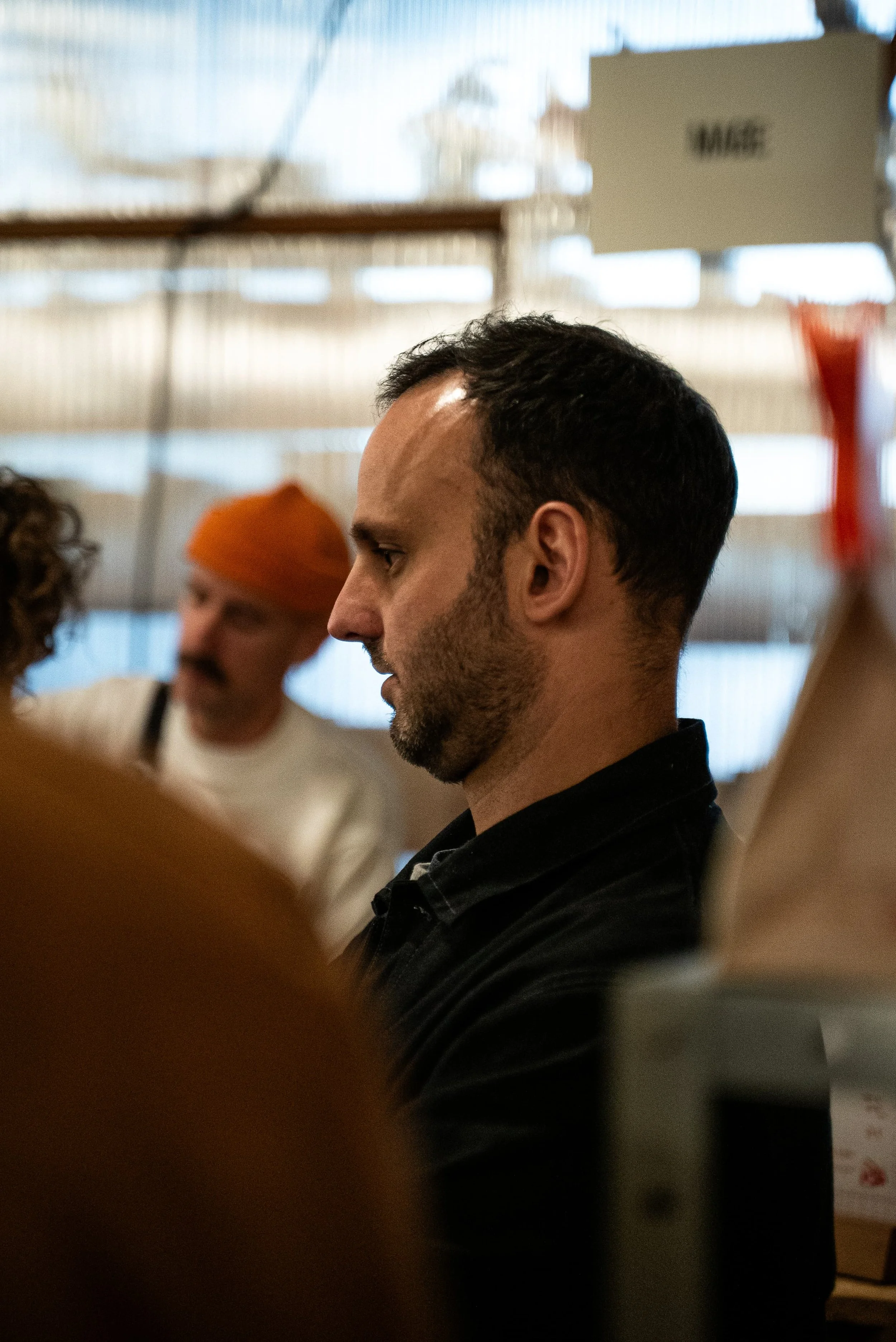 Un homme au profil sombre, portant une veste noire, dans un lieu intérieur, avec d'autres personnes en arrière-plan, dont une avec un chapeau orange, dans un environnement confus.