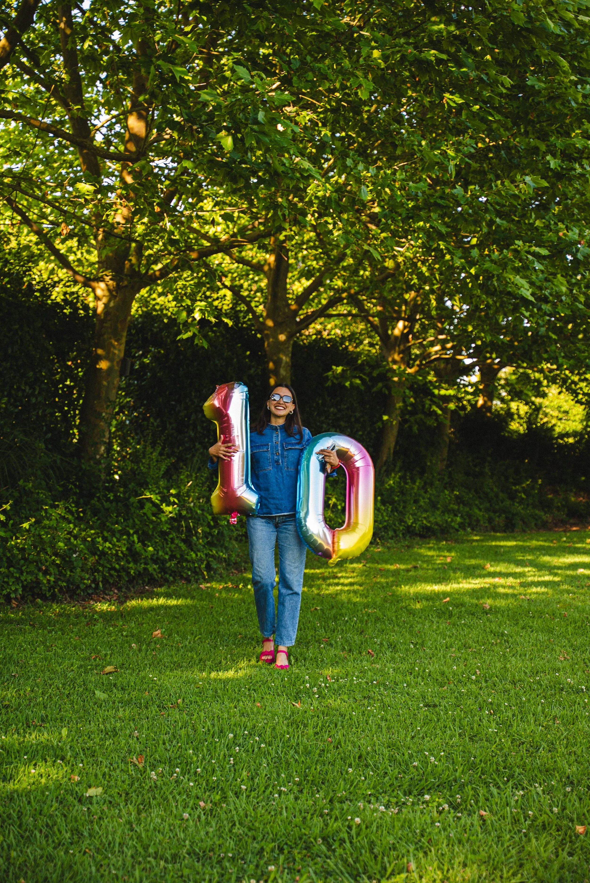 Une femme souriante portant des ballons numéros 1 et 0, debout sur une pelouse verte sous un arbre avec des feuilles vertes.