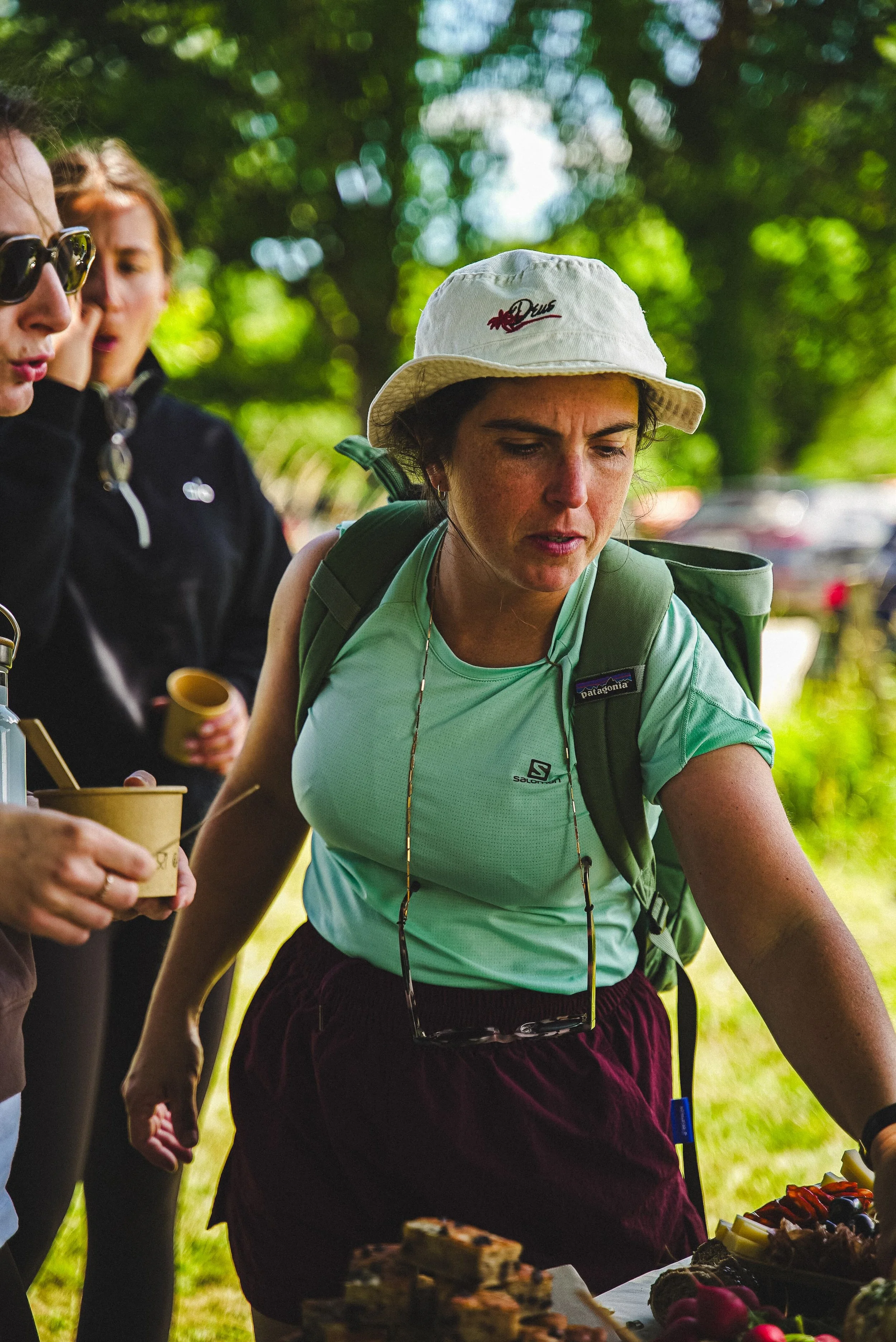 Groupe de personnes au pique-nique dans un parc, avec une femme portant un chapeau blanc, un sac à dos vert, et une table avec des desserts devant elle.