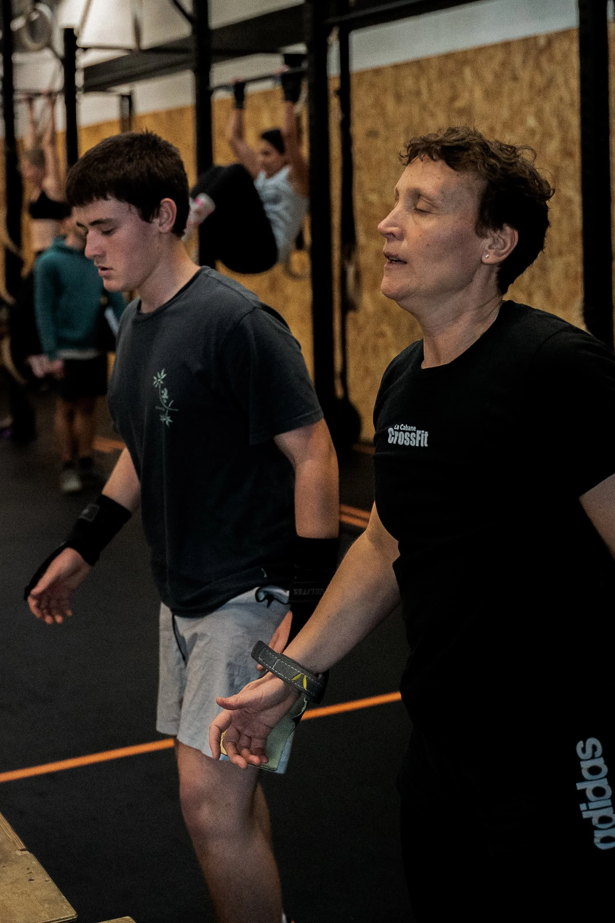 Deux personnes, une jeune homme et une femme plus âgée, participent à un entraînement avec des bandes de résistance dans une salle de sport CrossFit. L'ambiance est concentrée, et d'autres personnes en arrière-plan effectuent également des exercices.