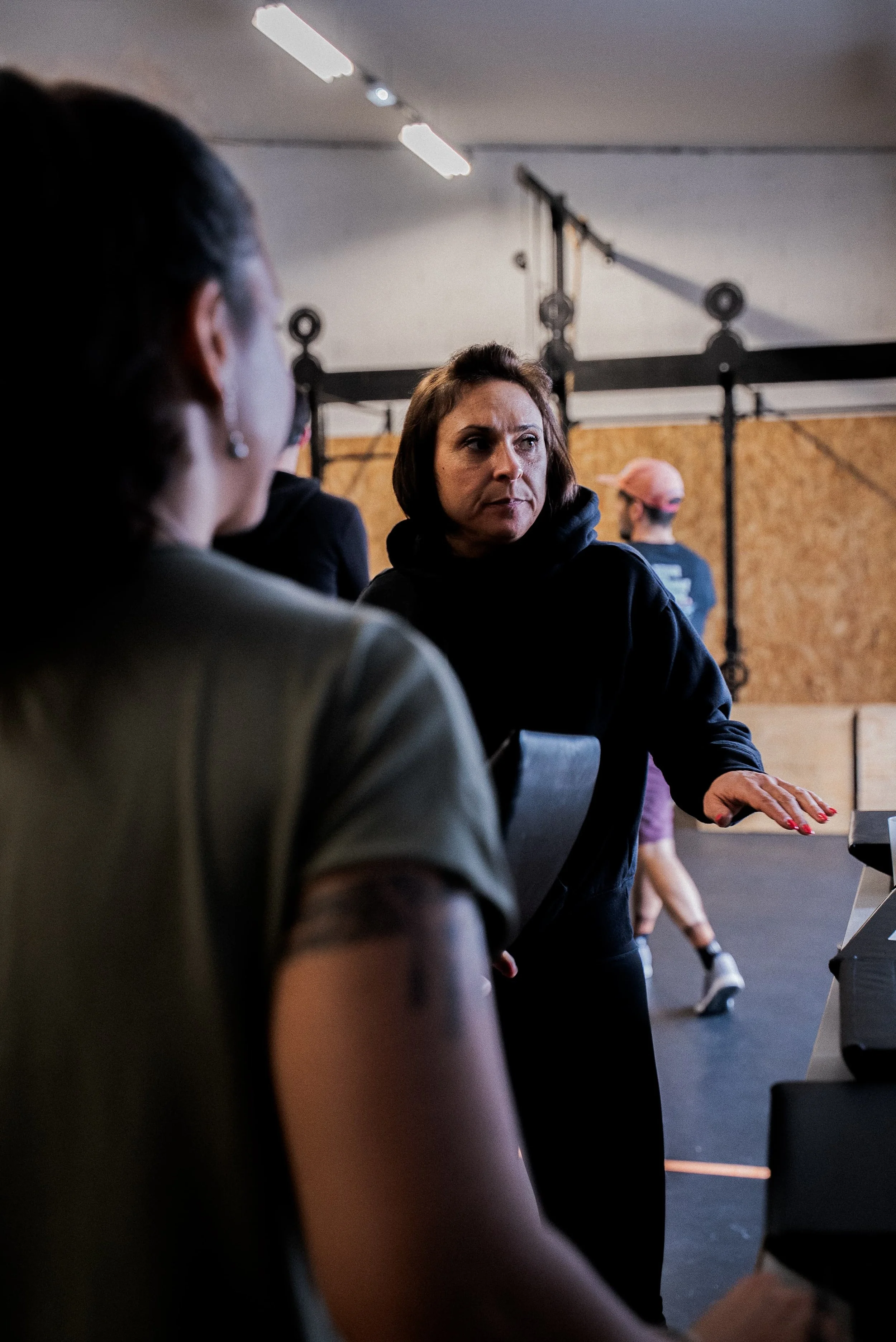 Une femme discute avec une autre personne dans une salle de sport ou un centre de fitness, avec un mur en bois et du matériel d'entraînement en arrière-plan.