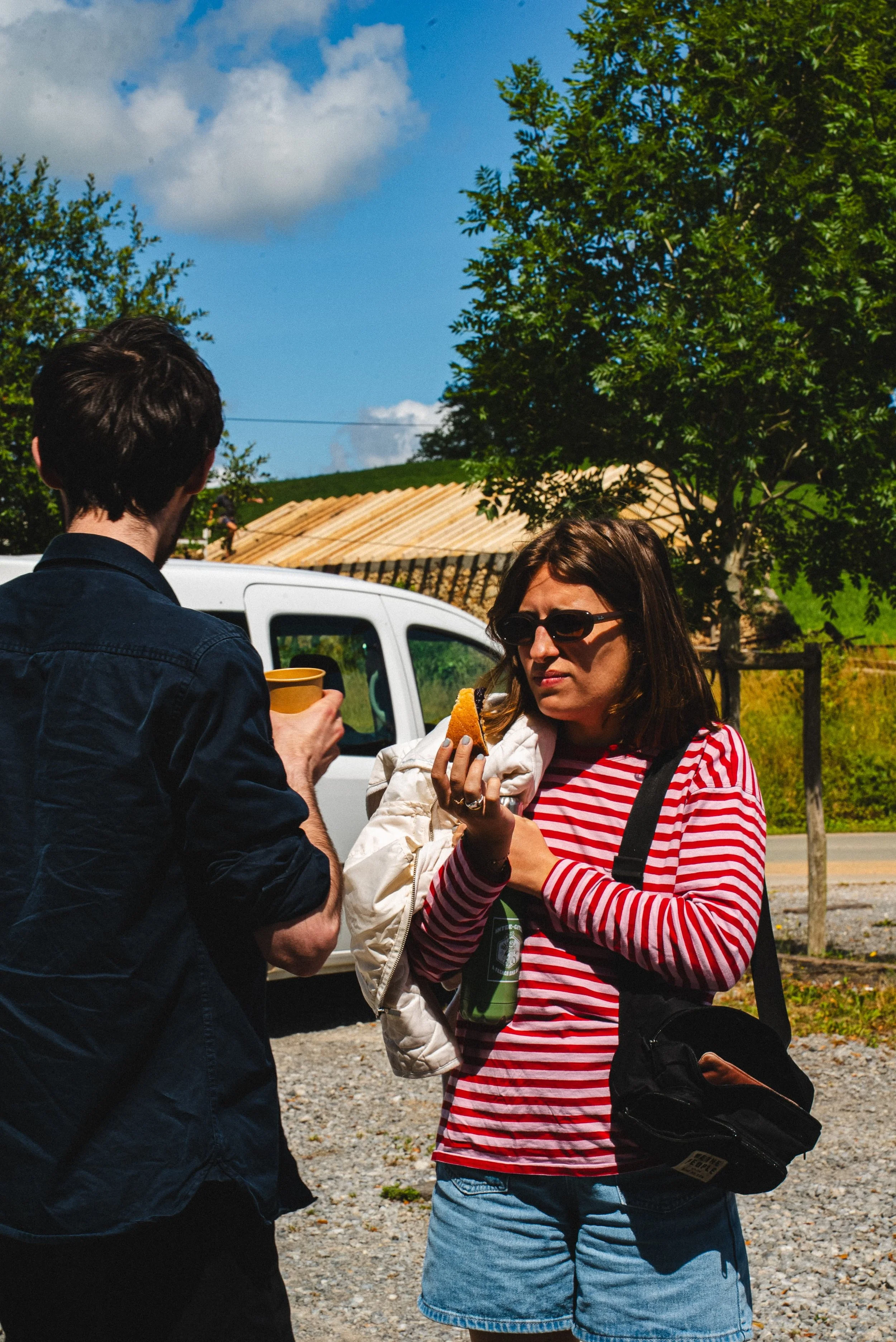Deux femmes discutent à l'extérieur, l'une portant des lunettes de soleil et une chemise à rayures rouges et blanches, l'autre en veston noir, devant une voiture blanche, avec des arbres et un bâtiment en arrière-plan.