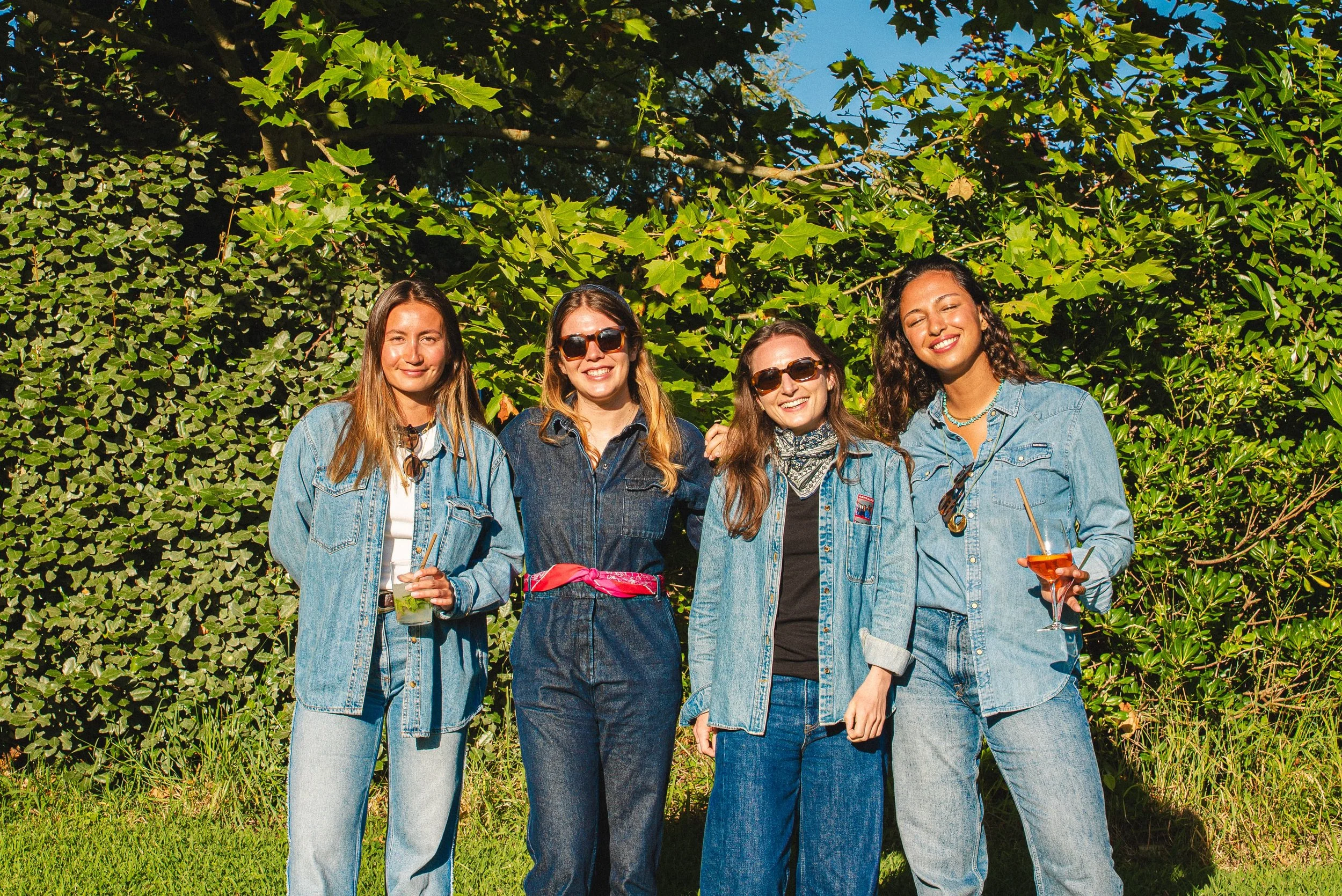 Quatre jeunes femmes portant des jeans décontractés, certaines avec des lunettes de soleil, souriantes, posant dehors devant un mur de feuilles vertes, tenant des boissons lors d'une journée ensoleillée.