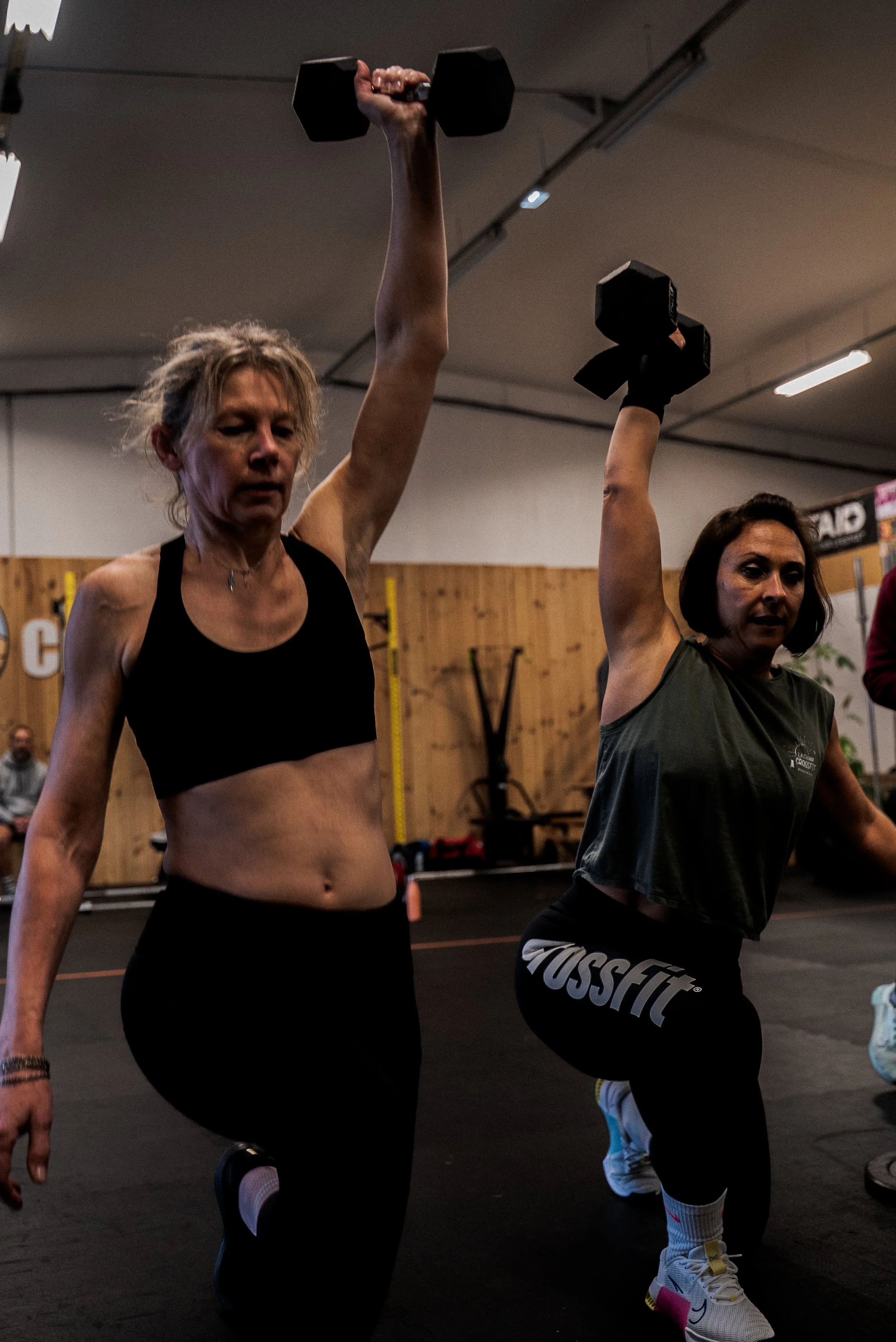 Deux femmes effectuants des exercices de musculation avec des haltères dans une salle de sport, l'une est en position debout et l'autre en position accroupie.