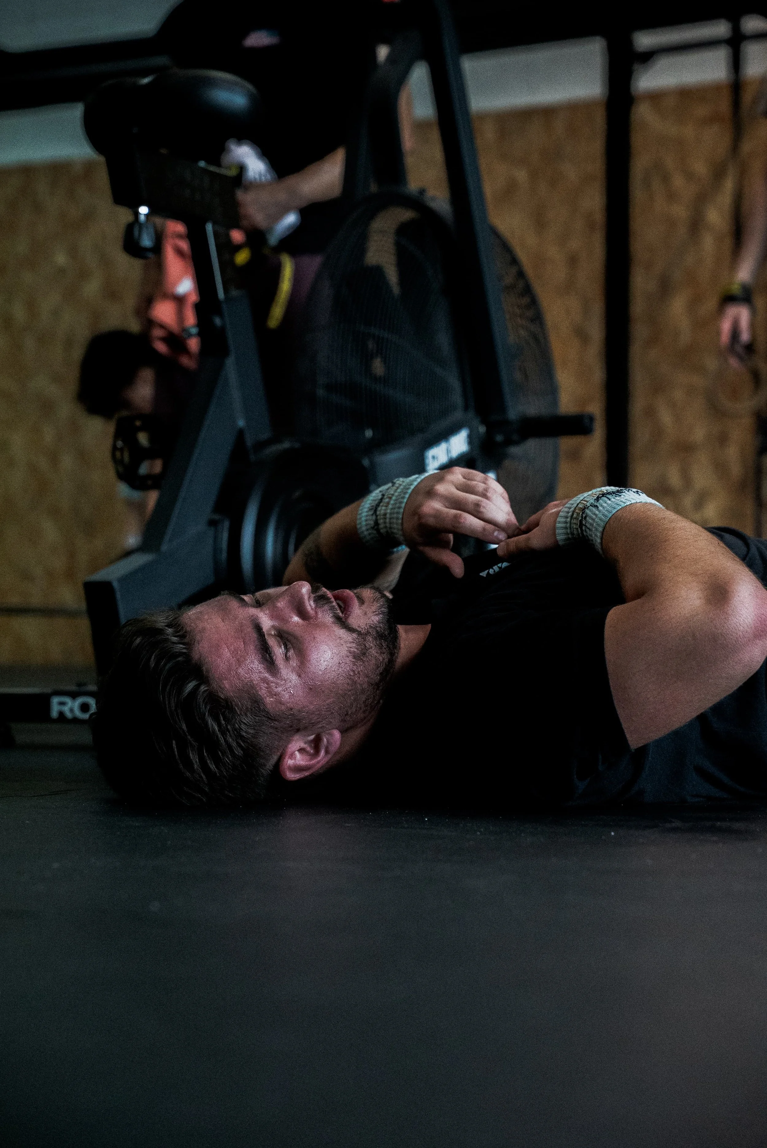 Un homme allongé sur le sol, tenant sa tête avec ses mains, à proximité d'une machine de musculation dans une salle de sport.