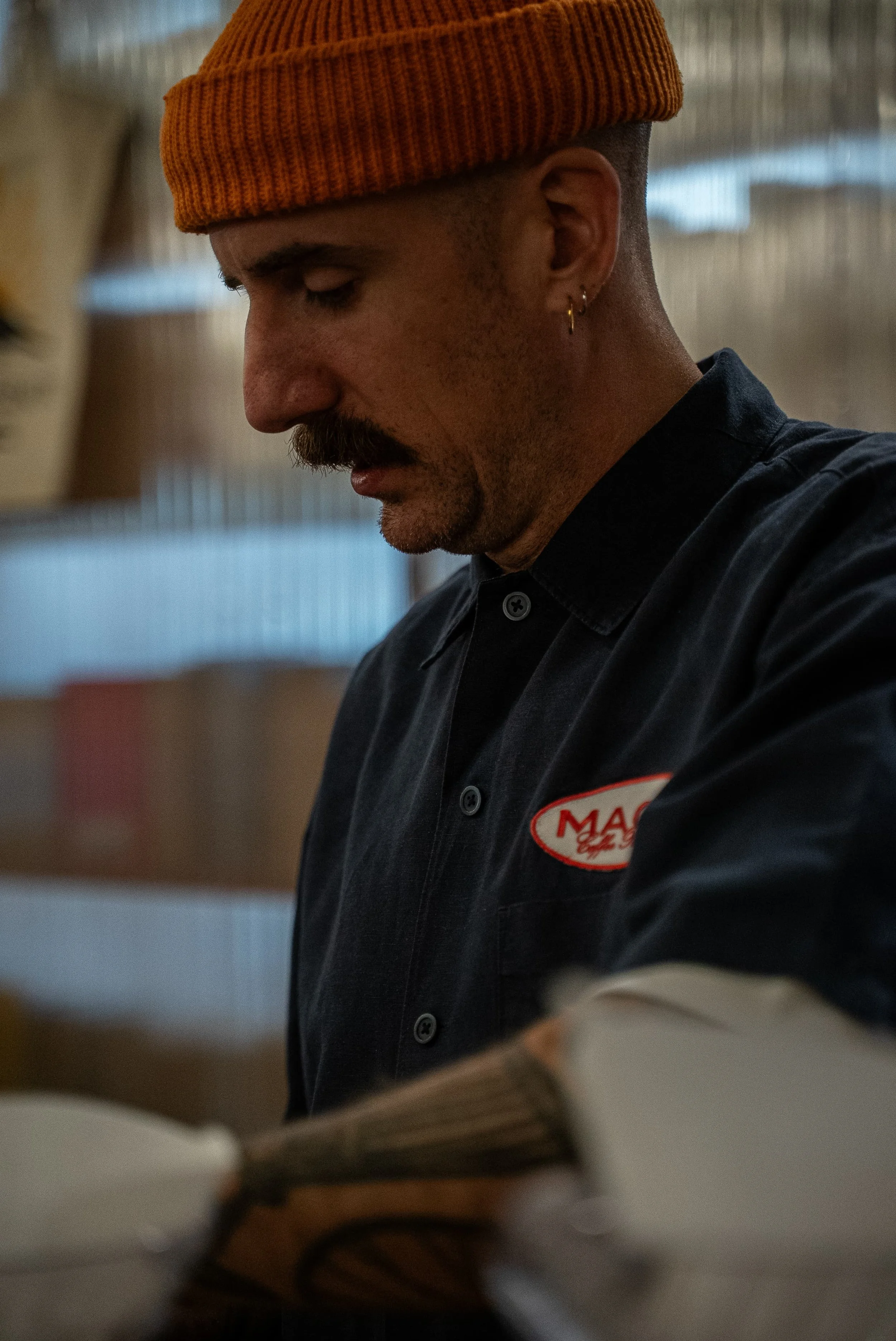 Portrait d'un homme portant un bonnet orange, une moustache, une barbe légère, avec une boucle d'oreille, en uniforme noir avec un badge 'MAG' sur la poitrine, en train de lire un livre dans un environnement intérieur.