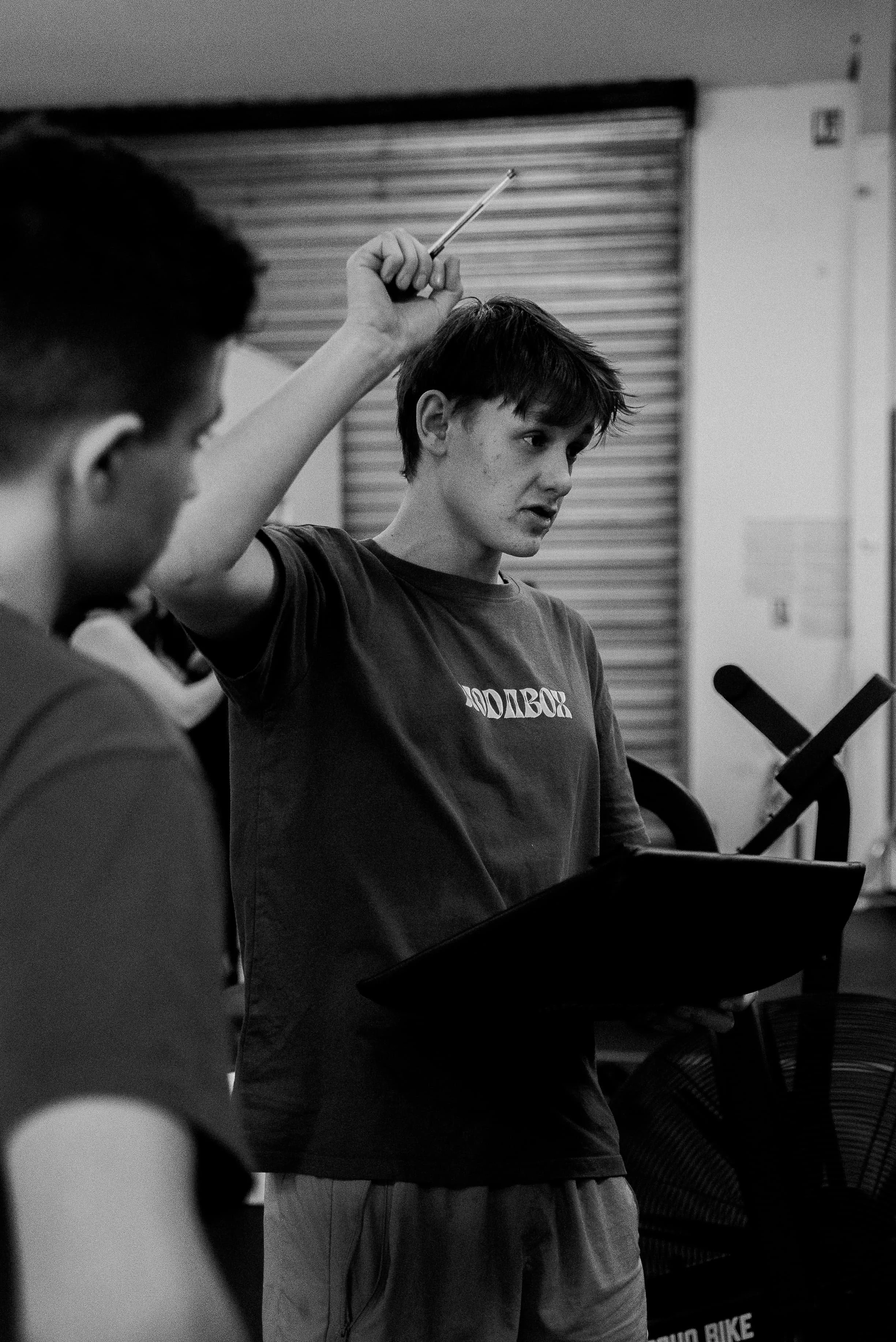 Deux jeunes garçons en train d'apprendre ou de jouer de la musique dans une salle intérieure, l'un d'eux tient un bâton ou un pinceau et semble chanter ou parler, tandis que l'autre est concentré sur quelque chose en face.
