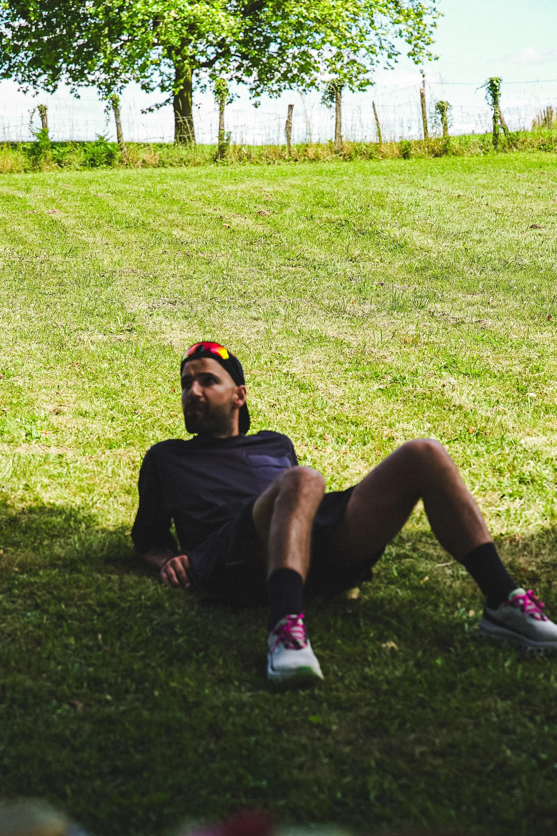 Jeune homme allongé sur l'herbe dans un parc, portant un t-shirt noir, un short noir, des chaussures de sport colorées, et une casquette noire avec un logo coloré, sous un arbre au soleil.