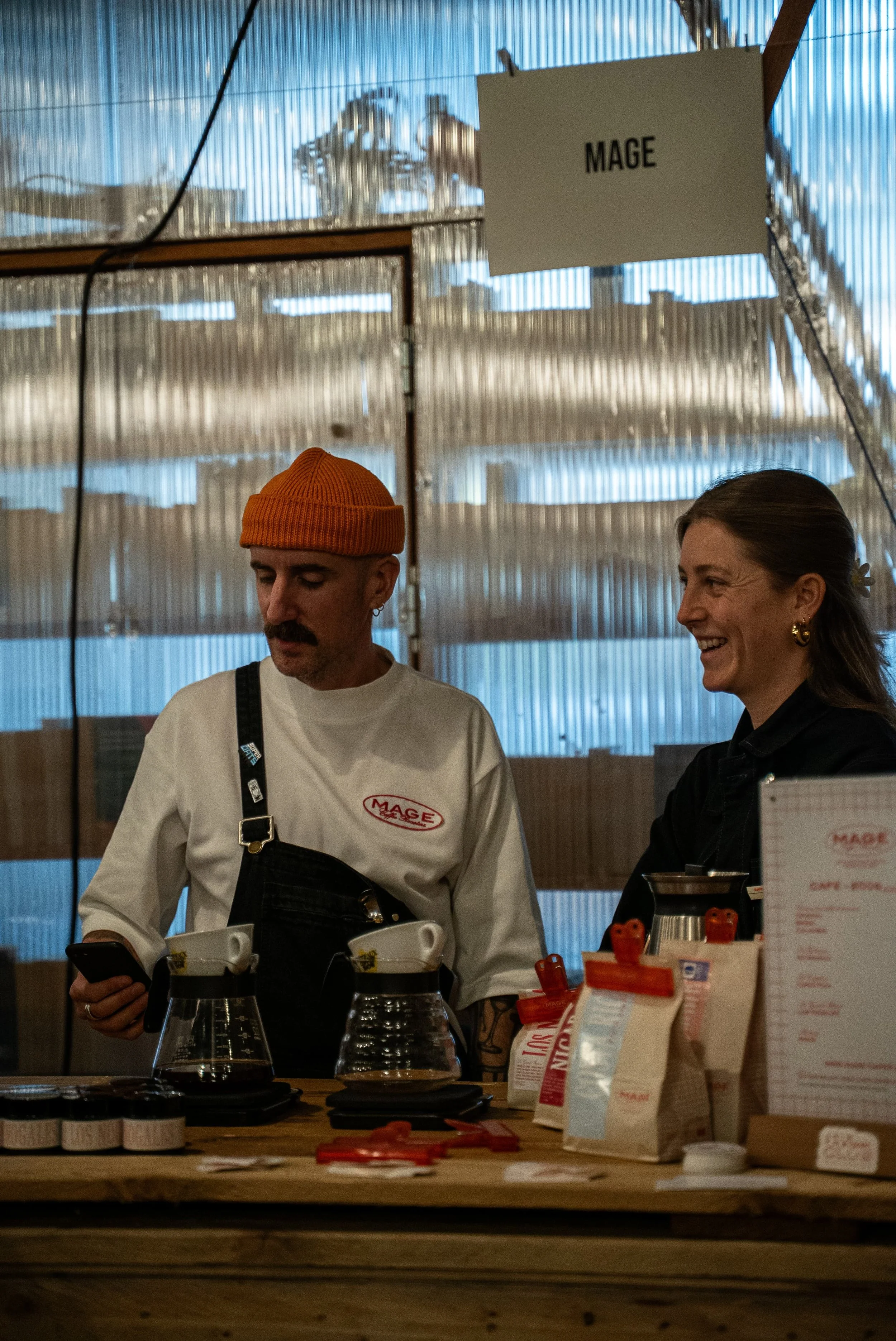Deux personnes derrière un comptoir de café, un homme avec un bonnet orange et un tatouage sur le bras, une femme souriante. Signale « MAGE » suspendu au plafond.