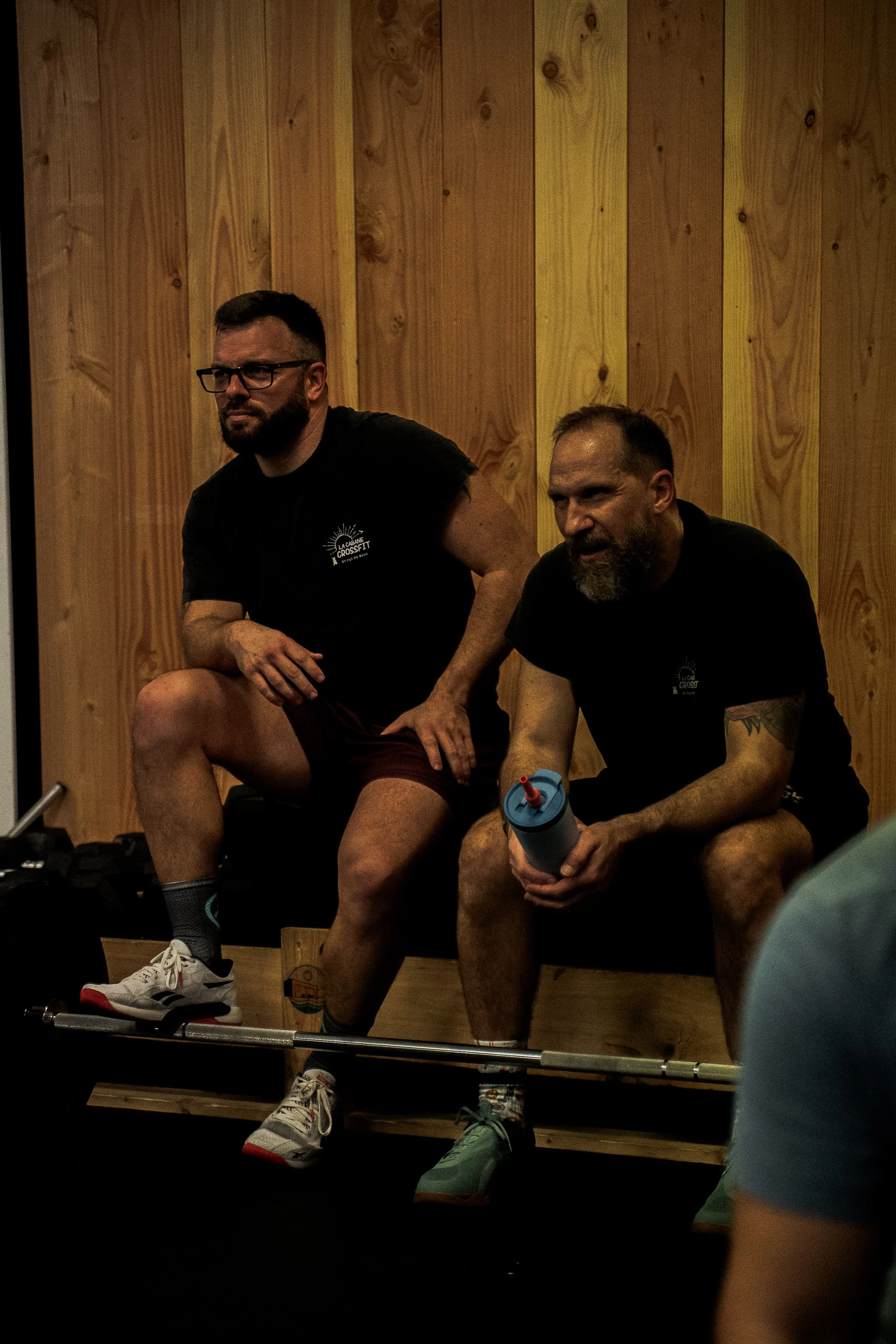 Deux hommes s'entraînent dans une salle de sport en bois, l'un tenant une bouteille d'eau et l'autre utilisant un équipement de musculation, tous deux en vêtements de sport noirs.