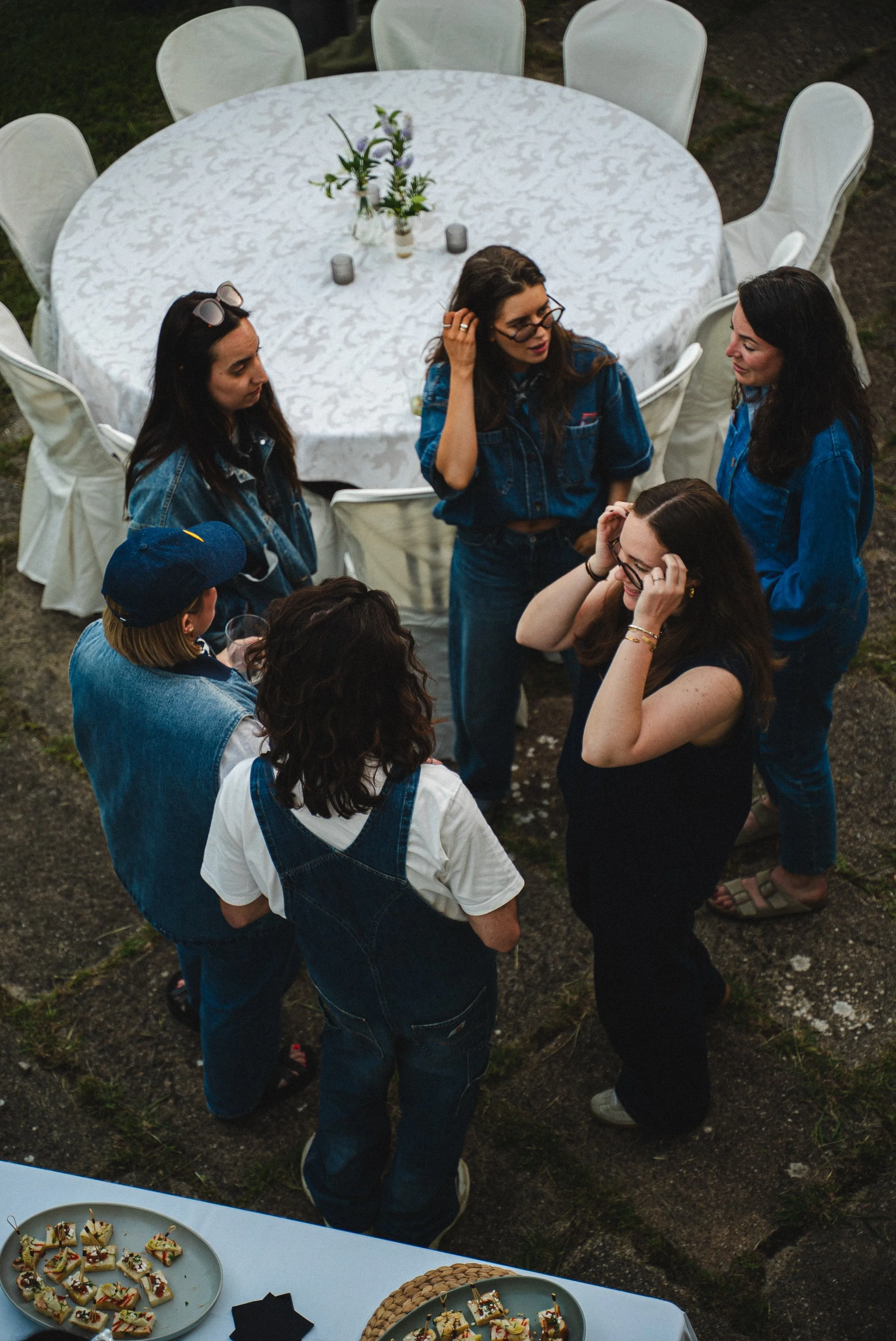 Un groupe de jeunes femmes discutant lors d'une fête en extérieur, avec une table ronde couverte d'une nappe blanche et des plats de snacks en premier plan.