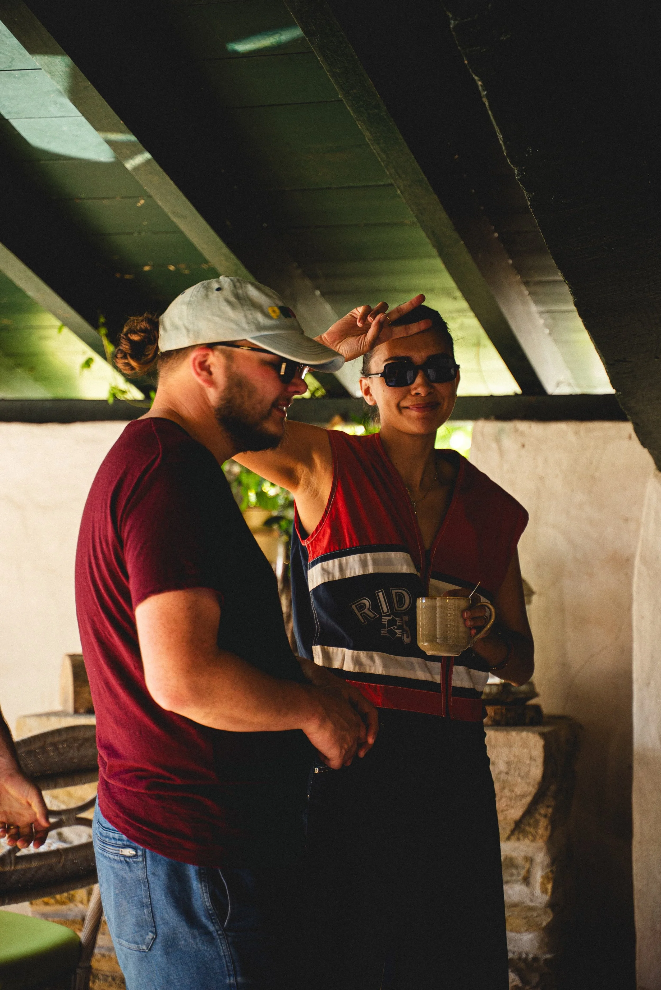 Deux personnes discutent dans une ambiance décontractée, l'une d'elles tenant une tasse, sous un plafond en bois. L'homme porte une casquette et des lunettes de soleil, la femme porte également des lunettes de soleil et un vêtement coloré.