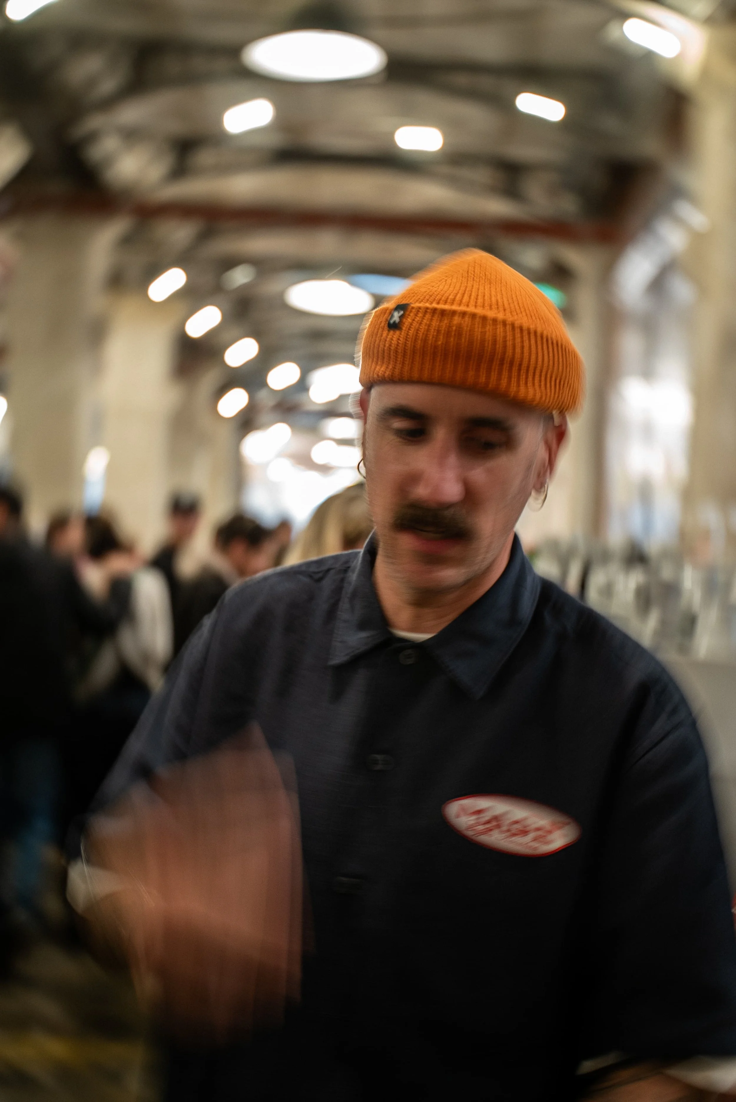 Un homme portant un bonnet orange et une chemise noire, dans une ambiance de restaurant ou café, entouré de plusieurs personnes en arrière-plan.