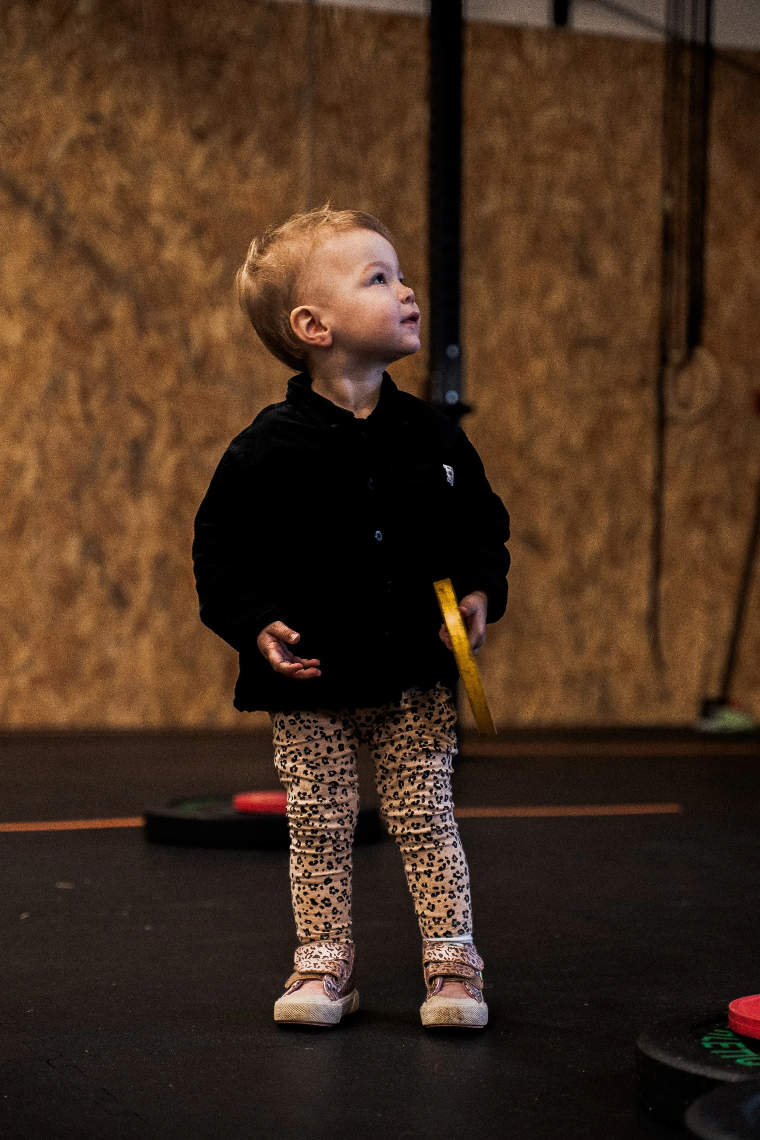 Un jeune enfant debout sur un tapis de sol, tenant un objet jaune dans une main, dans une salle avec un mur en bois en arrière-plan.