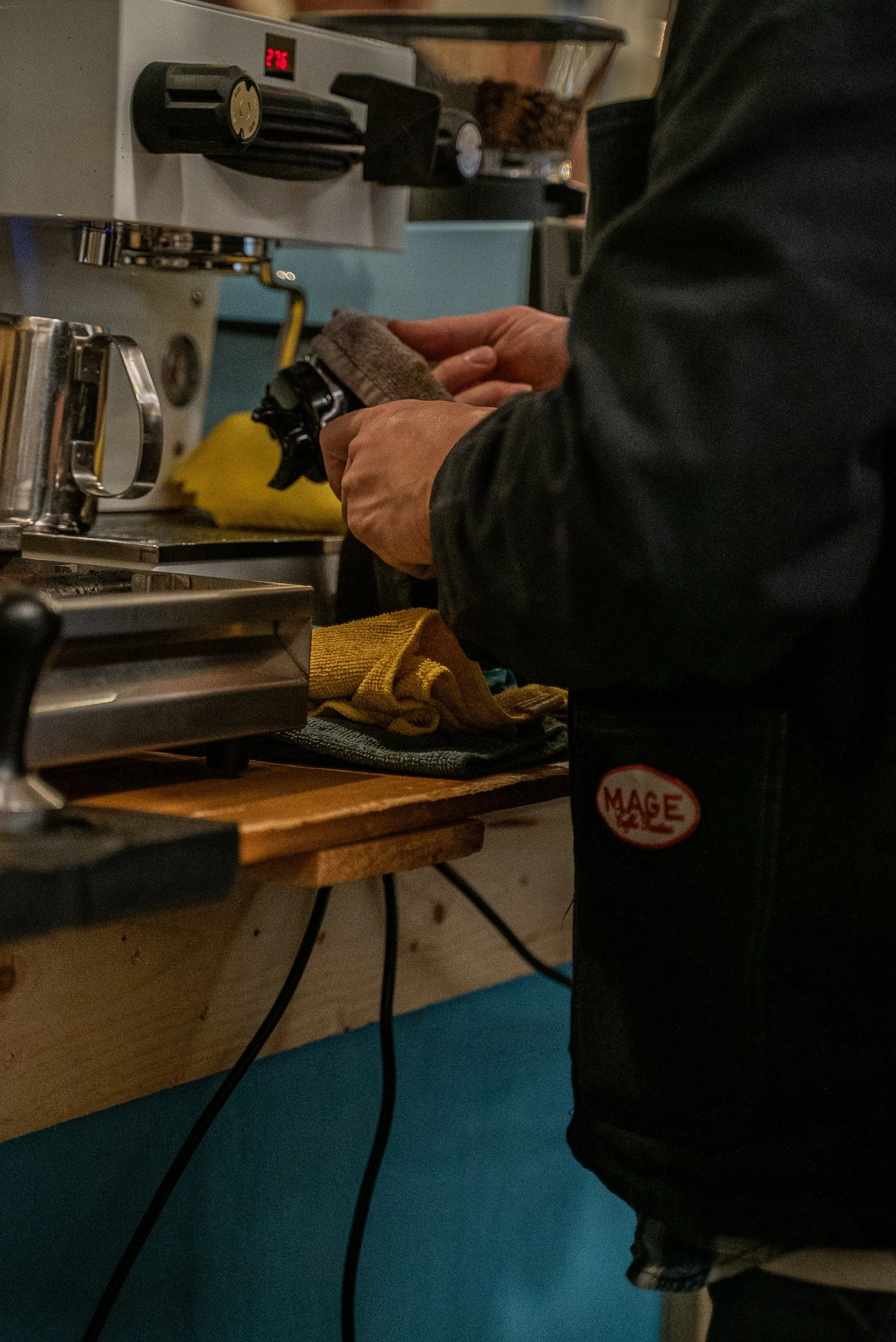 Une personne en train de préparer une boisson dans un café, avec une machine à café en arrière-plan affichant une température de 27,5°C.
