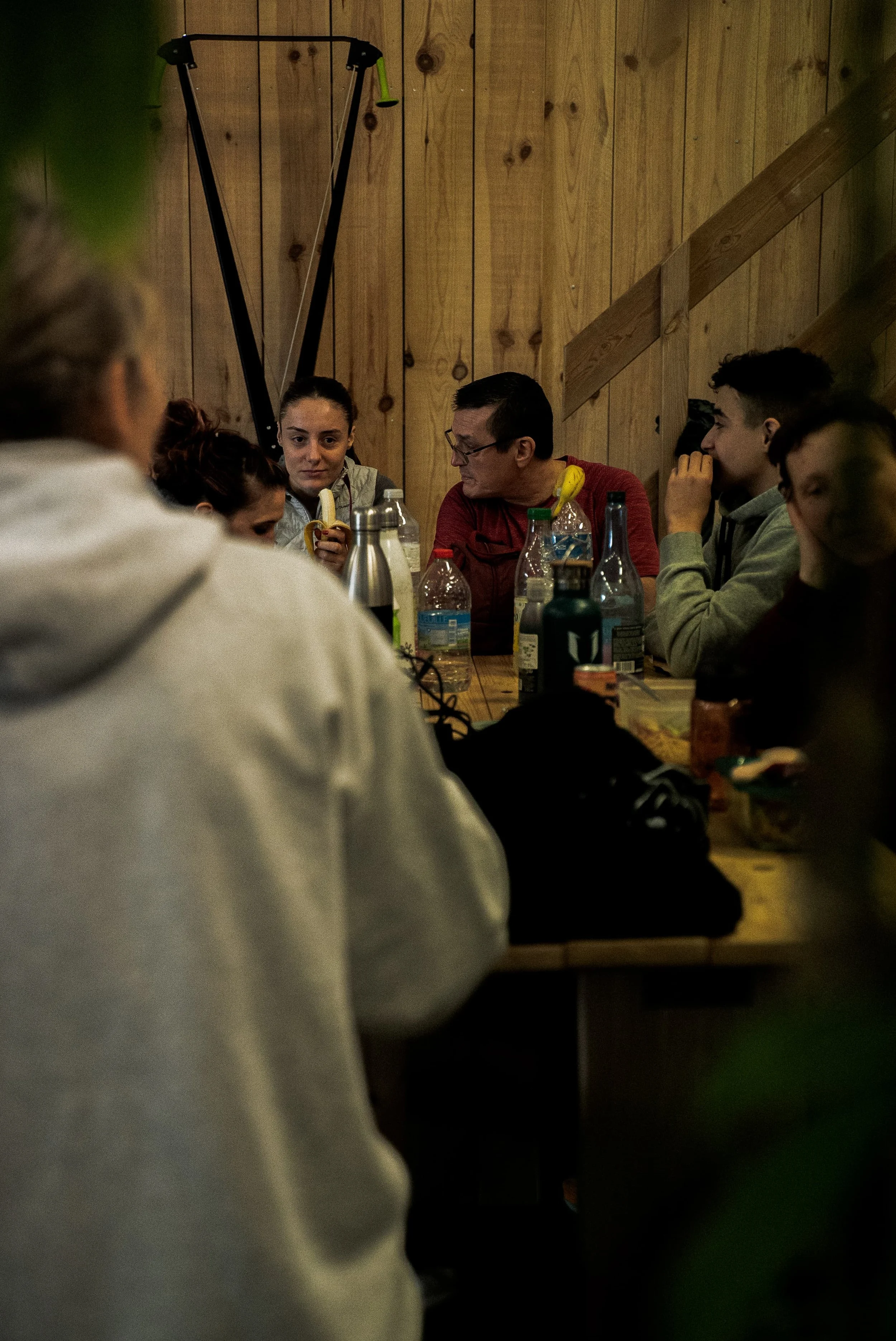 Groupe de personnes assises autour d'une table en bois dans une pièce avec un mur en bois visible, dégustant ou discutant.