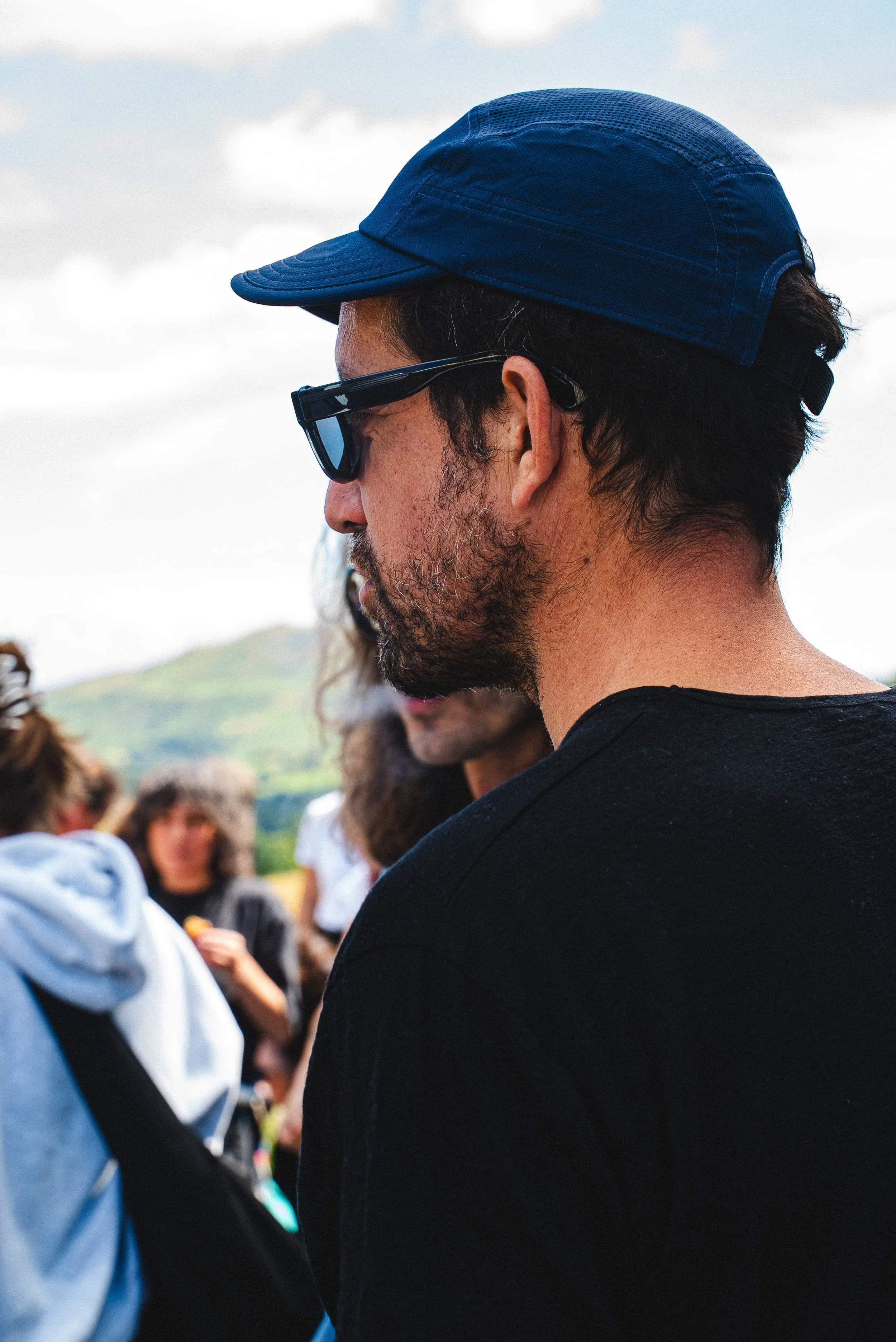 Un homme portant une casquette bleue, des lunettes de soleil noires, et une barbe, entouré d'autres personnes dans un environnement extérieur.