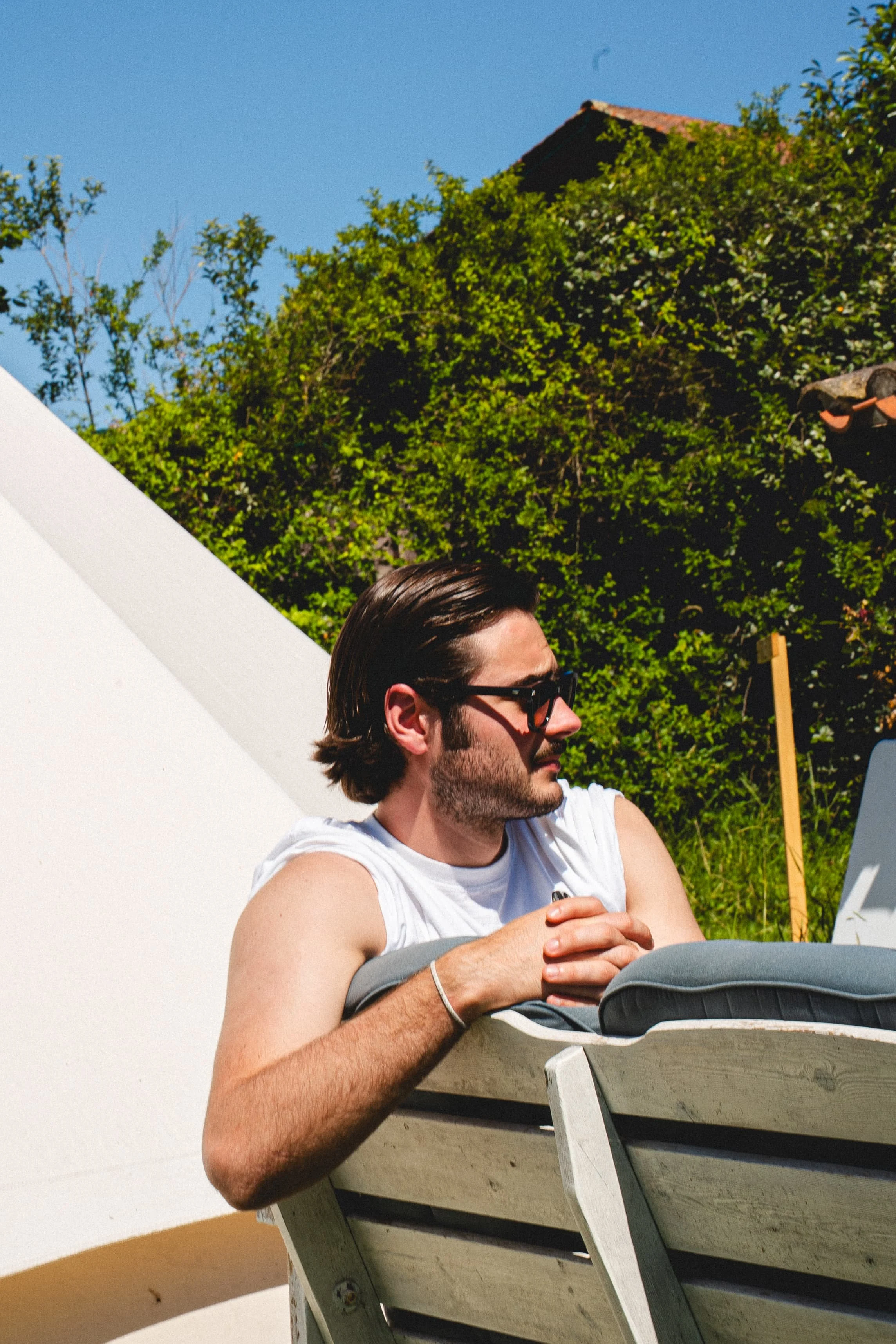 Un homme avec des cheveux bruns, portant des lunettes noires, s'appuie sur un banc en bois blanc, en extérieur, sous un ciel bleu avec des arbres verts en arrière-plan.