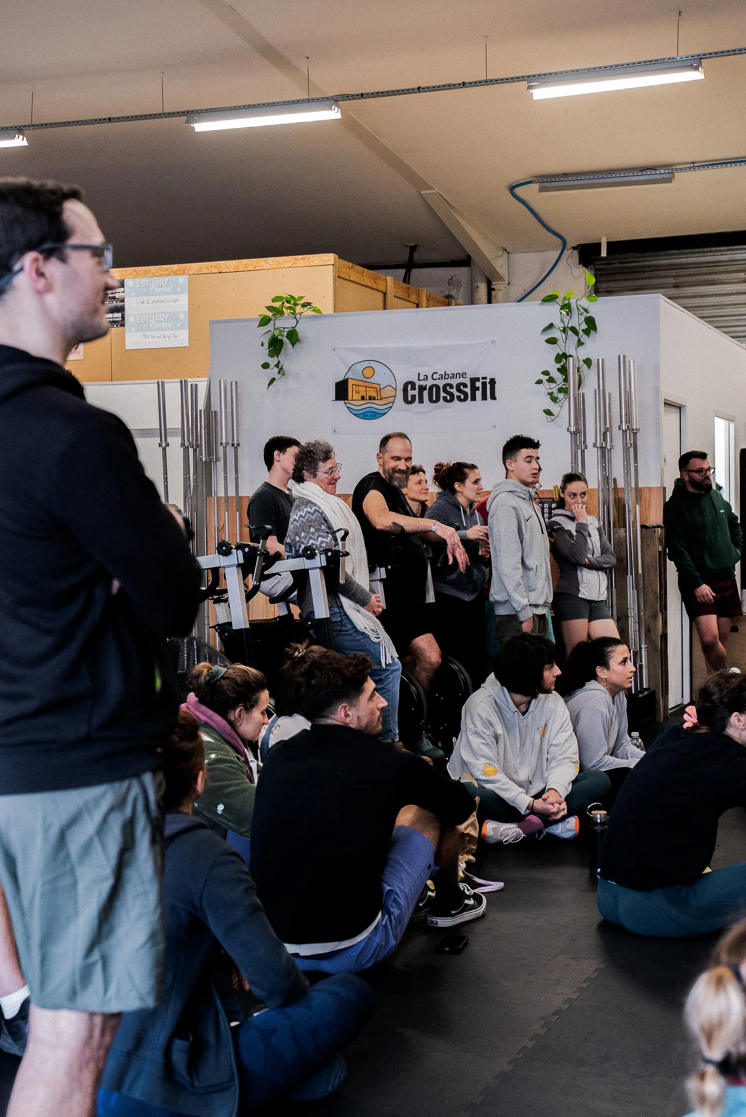 Groupe de personnes assises en méditation ou en attention dans une salle de CrossFit appelée La Cabane, avec un mur portant un logo et des plantes vertes.