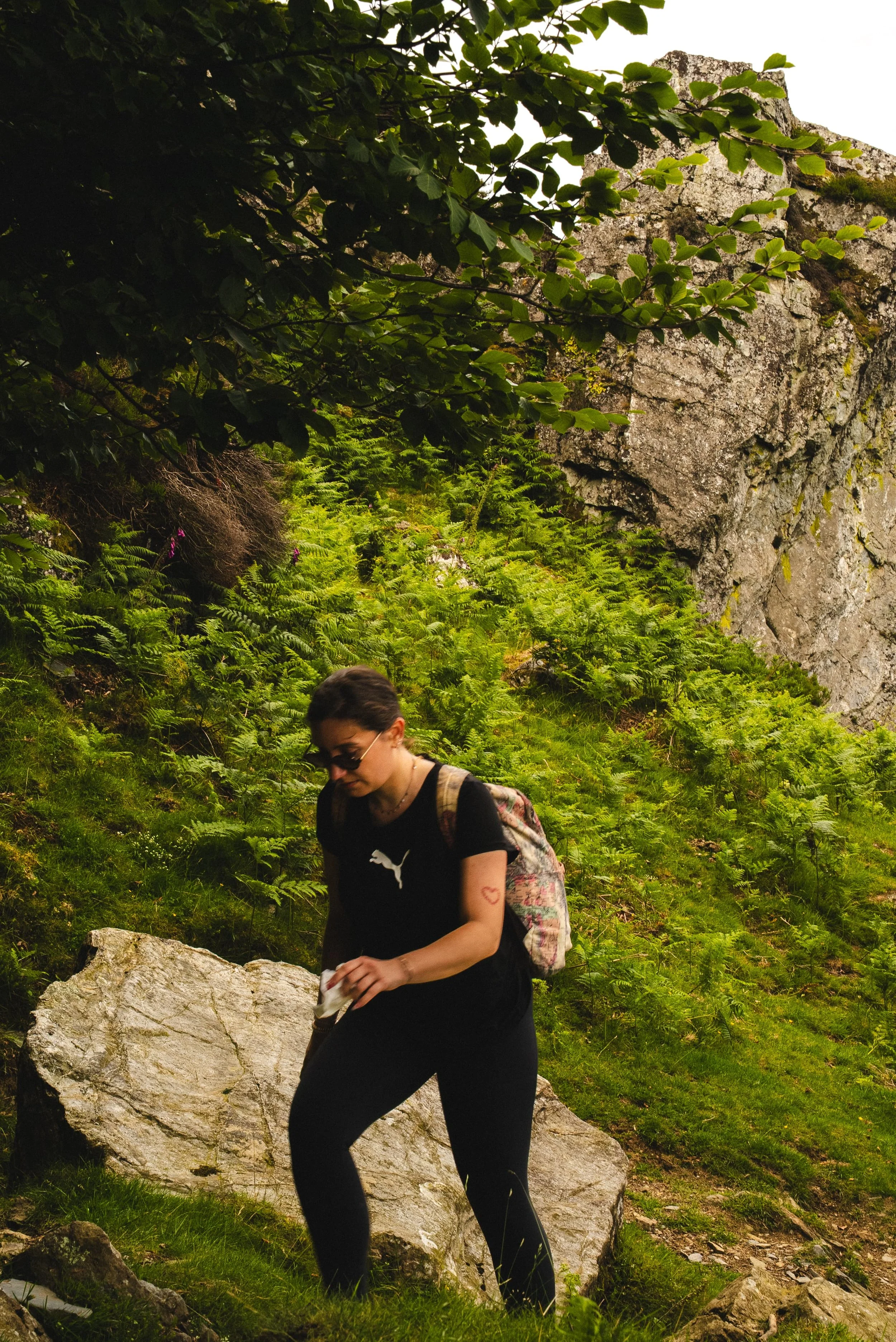 Jeune femme portant un t-shirt noir avec un logo blanc, des lunettes de soleil, un sac à dos, en train de marcher dans une nature verdoyante avec des rochers et des arbustes.