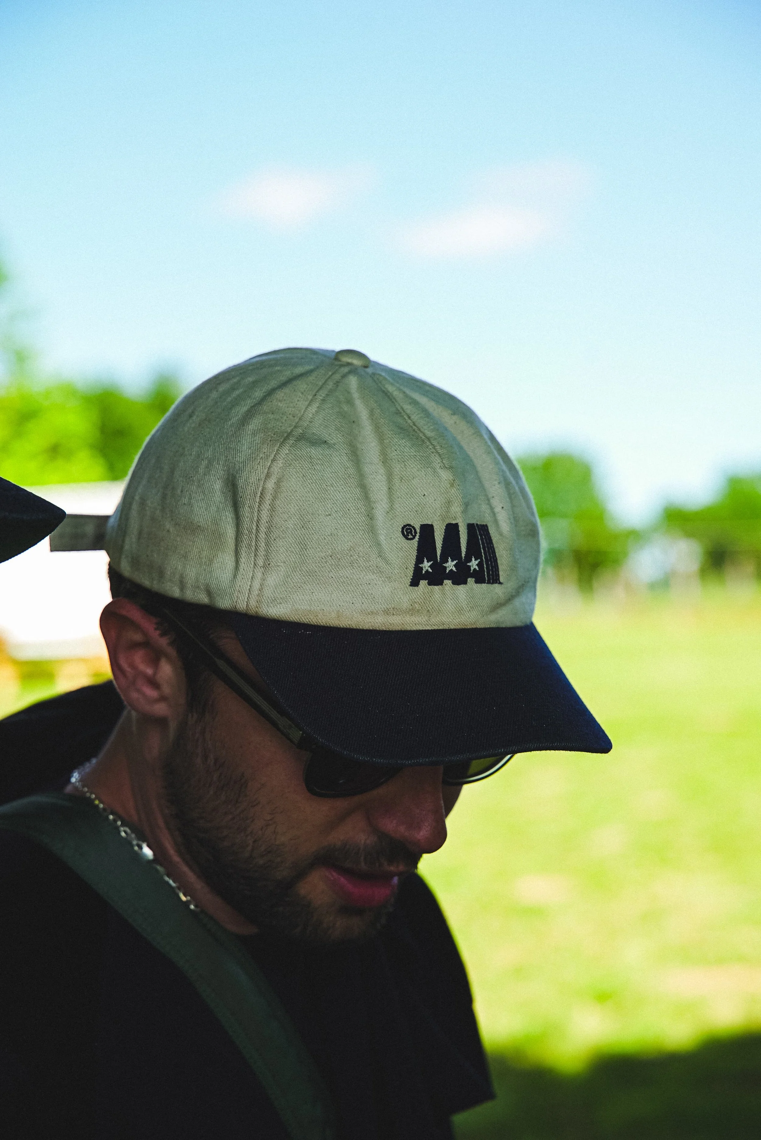 Un homme portant une casquette beige avec le logo 'AAA', lunettes de soleil et collier, en extérieur avec un ciel bleu et des arbres en arrière-plan.