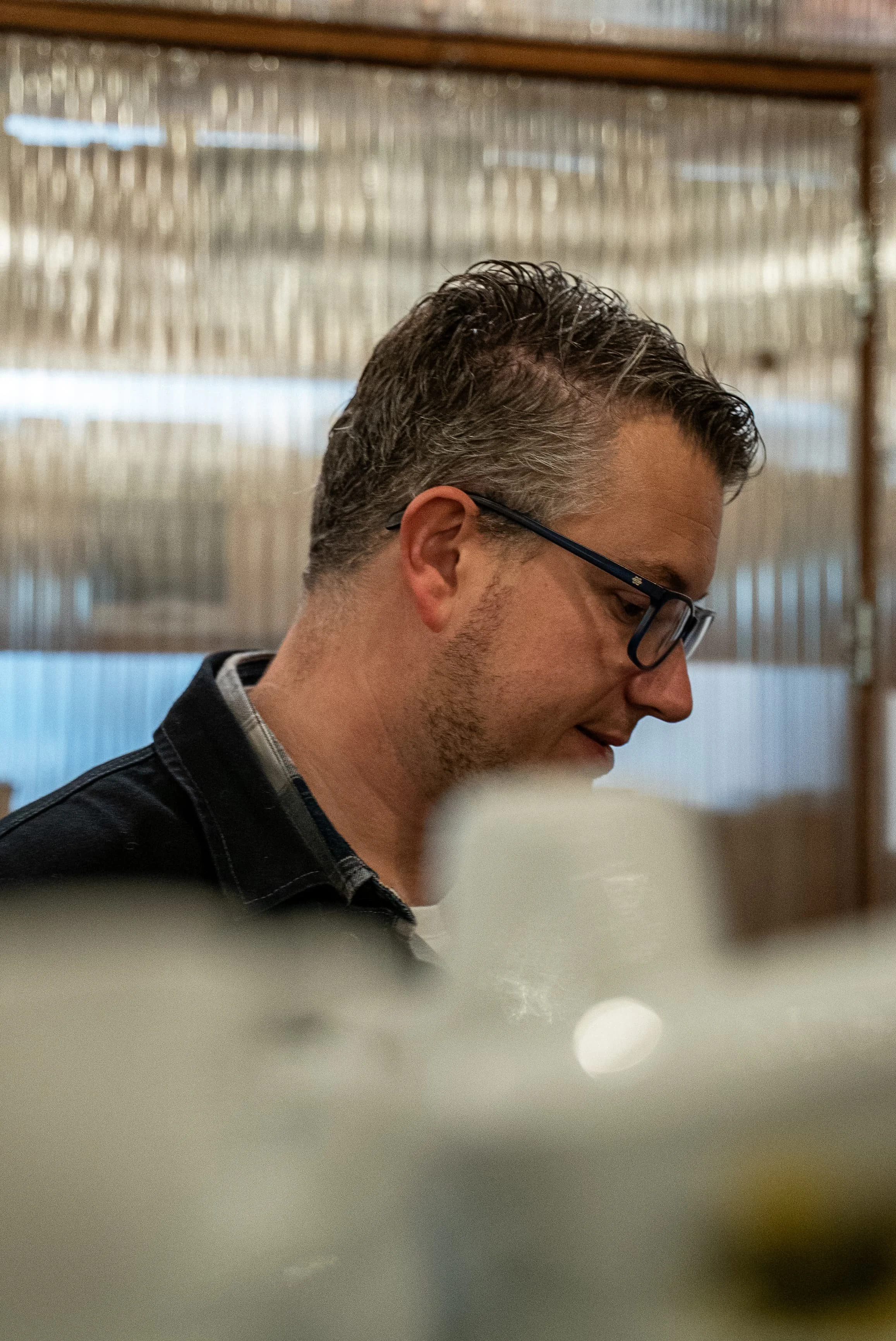 Un homme avec des cheveux courts et bouclés, portant des lunettes et un pull à carreaux, vu en profil, souriant, dans un intérieur avec une paroi en bois et un mur en verre à l'arrière.