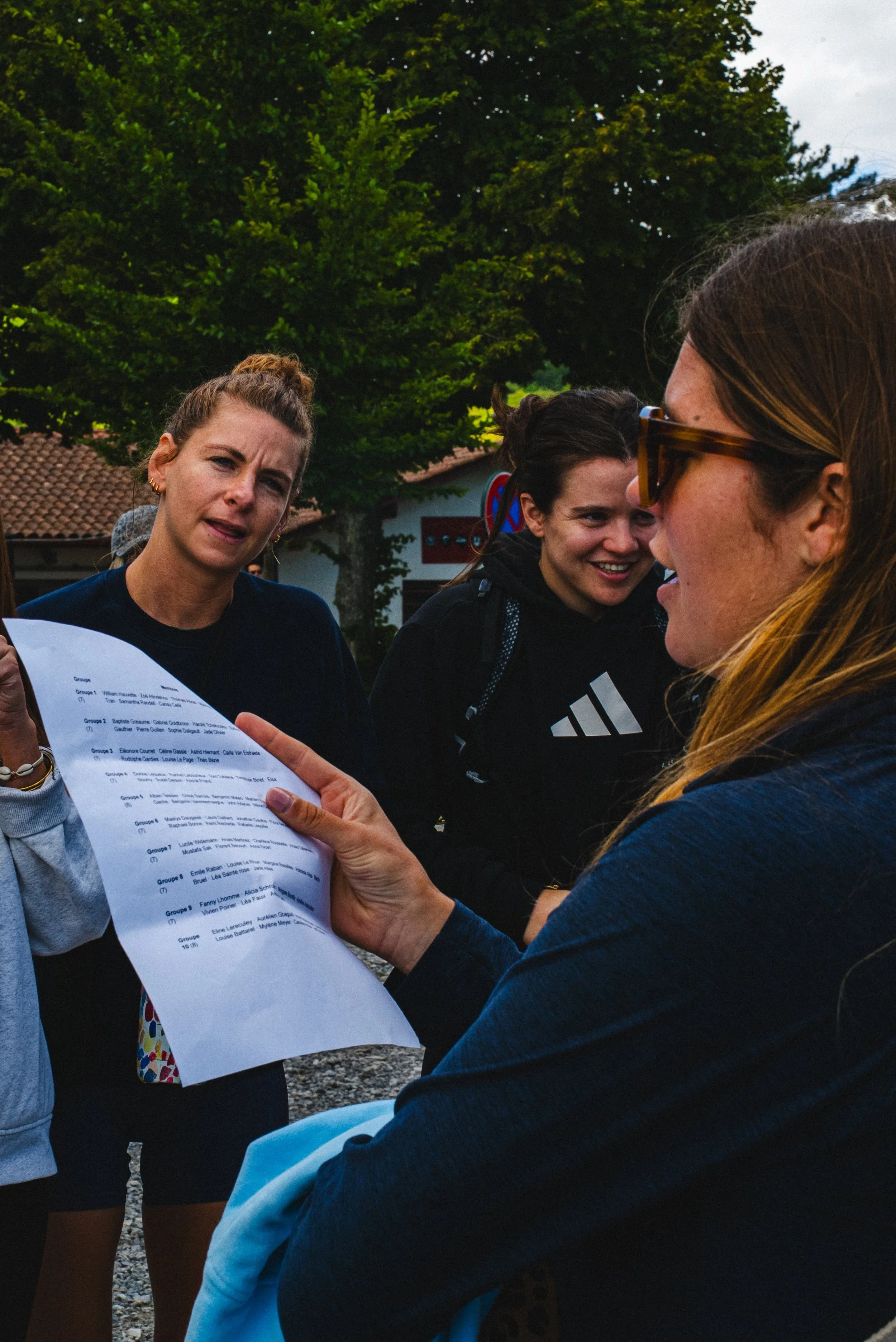 Groupe de jeunes femmes discutant en extérieur, une tenant un document, avec des arbres en arrière-plan.