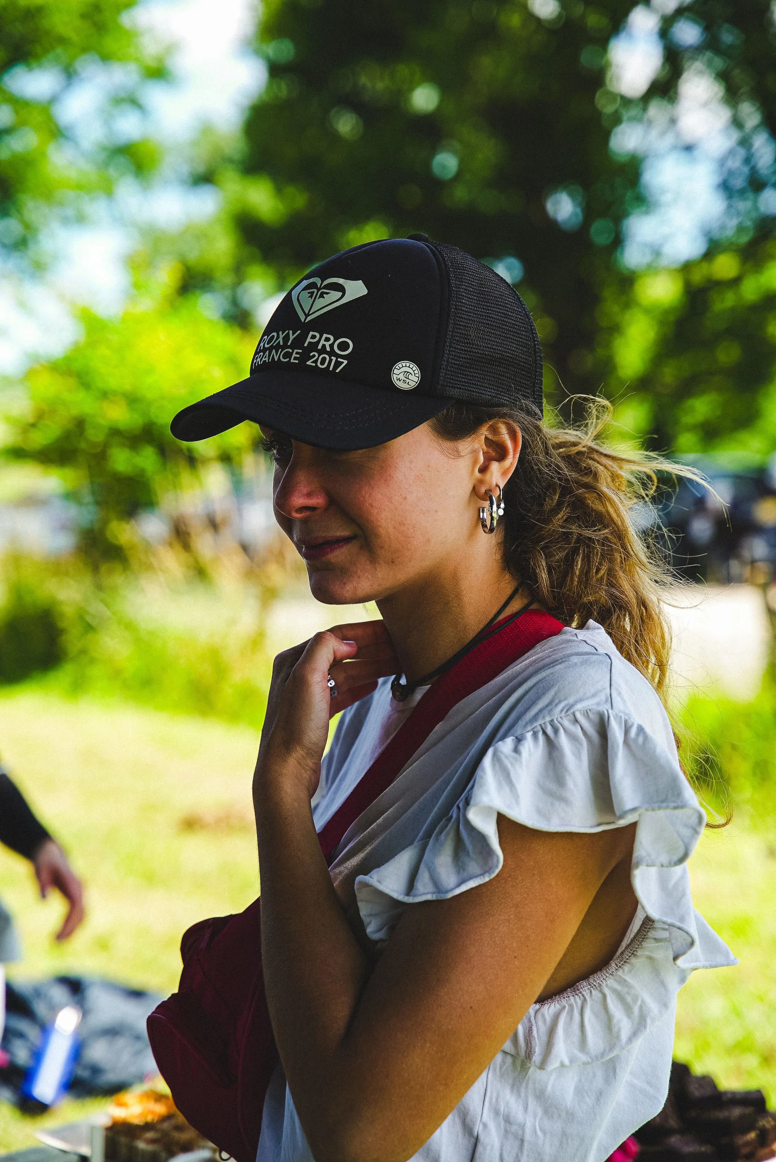 Une femme avec des boucles d'oreilles, portant un chapeau noir marqué "Roxy Pro France 2017" et une chemise blanche à manches courtes, se tenant dans un environnement extérieur avec des arbres en arrière-plan.
