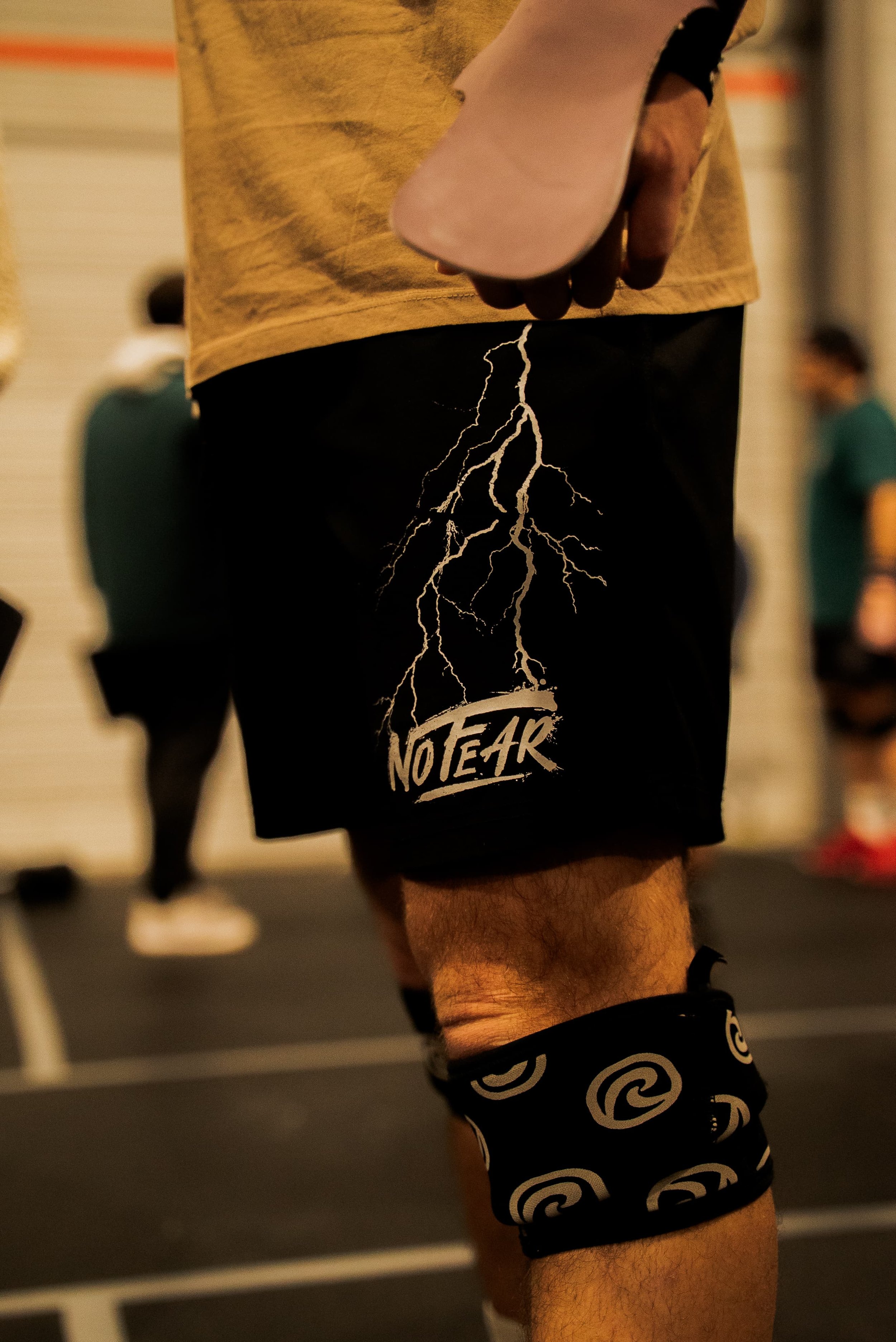 Une personne en tenue de sport portant un short noir avec un motif d'éclairs et le texte 'NO TEAR', équipée de brassards immaculés avec un motif en spirale, probablement dans une salle de sport ou lors d'une séance d'entraînement.