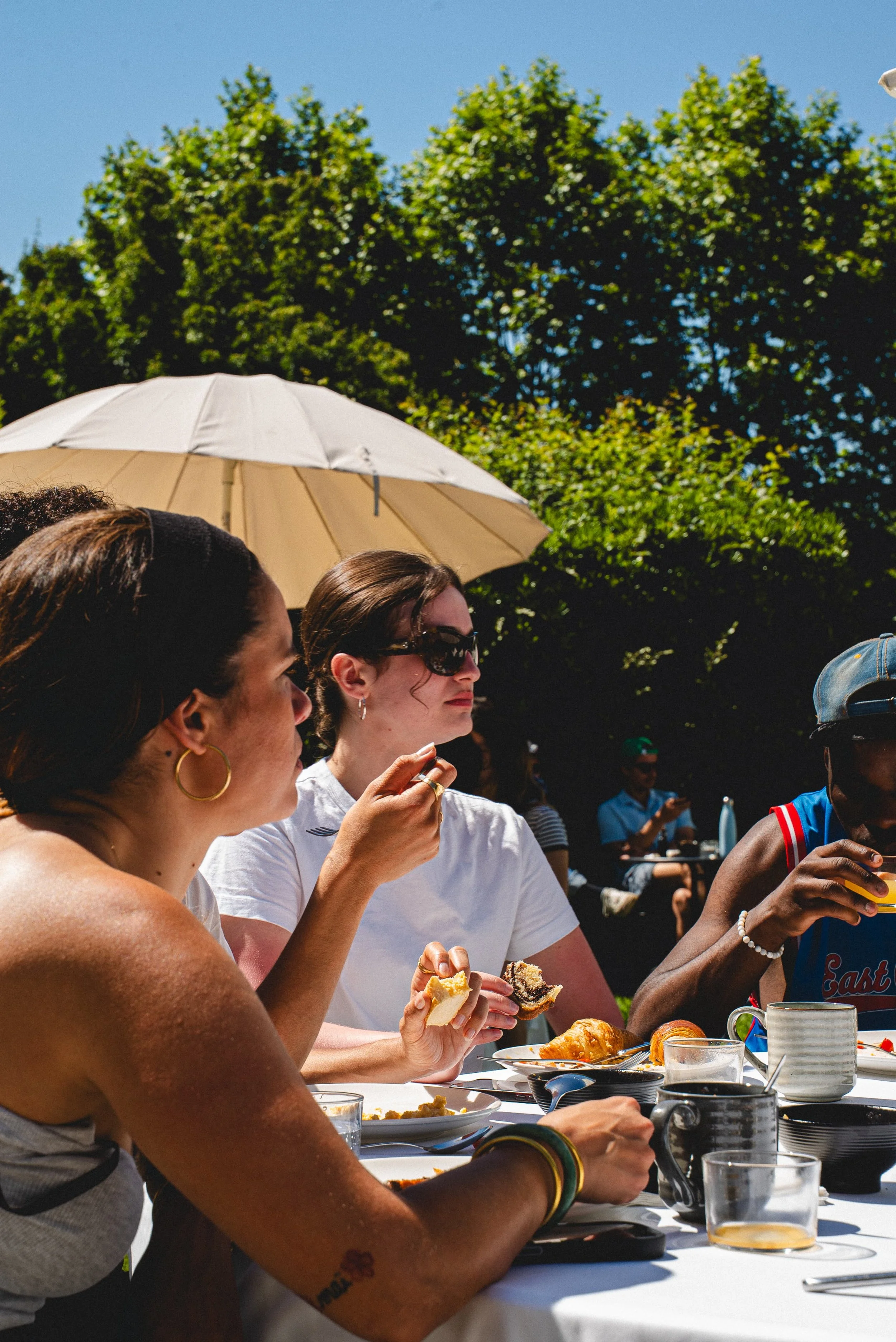 Groupe de personnes mangeant un repas en extérieur sous un parasol, avec des arbres en arrière-plan, par temps ensoleillé.
