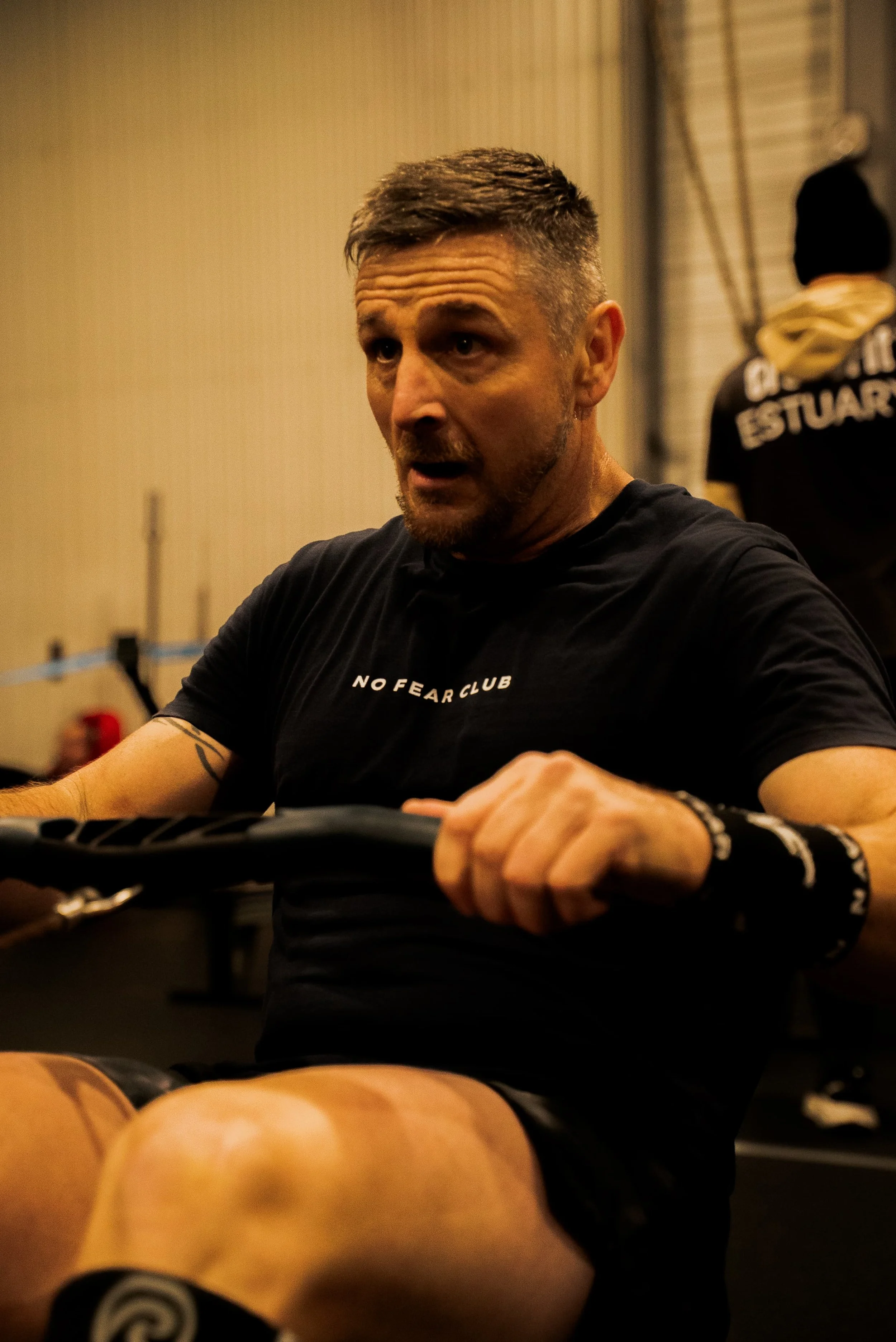 Un homme qui parle ou s'entraîne dans une salle de sport, portant un t-shirt noir avec l'inscription 'NO FEAR CLUB'.