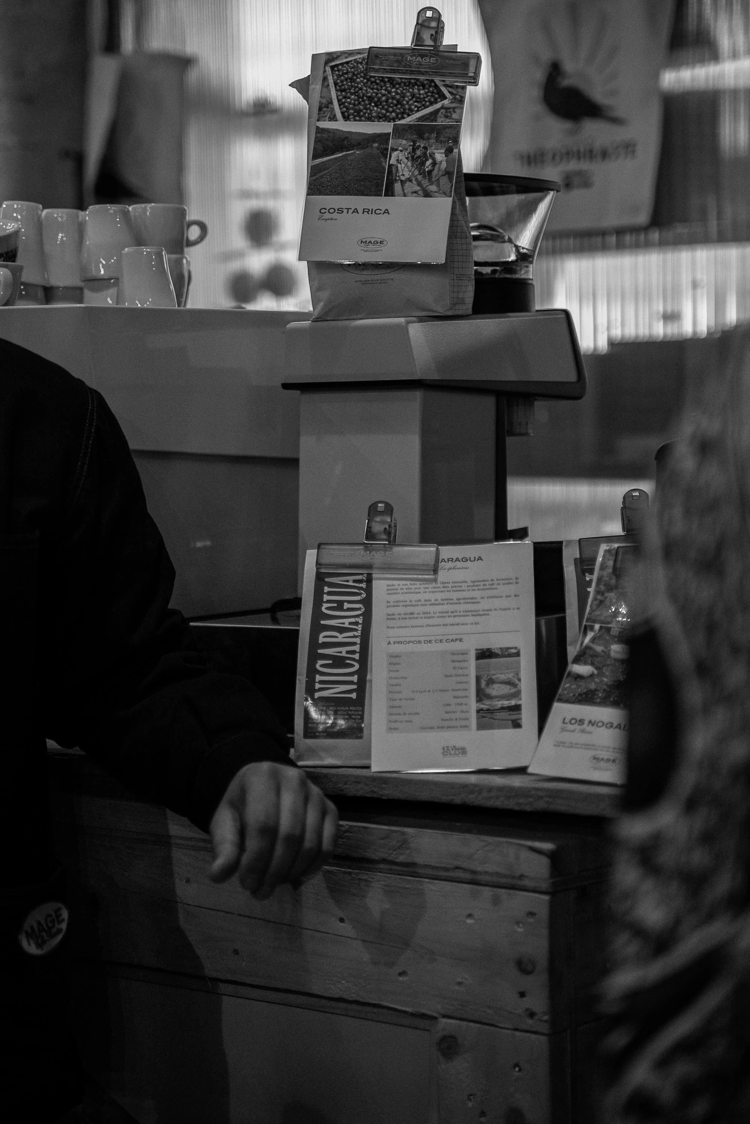 Une table en bois avec un homme qui tient une cigarette, des tasses blanches et des flyers ou menus affichés. Un sac de café Costa Rica et une machine à café sont visibles en arrière-plan.
