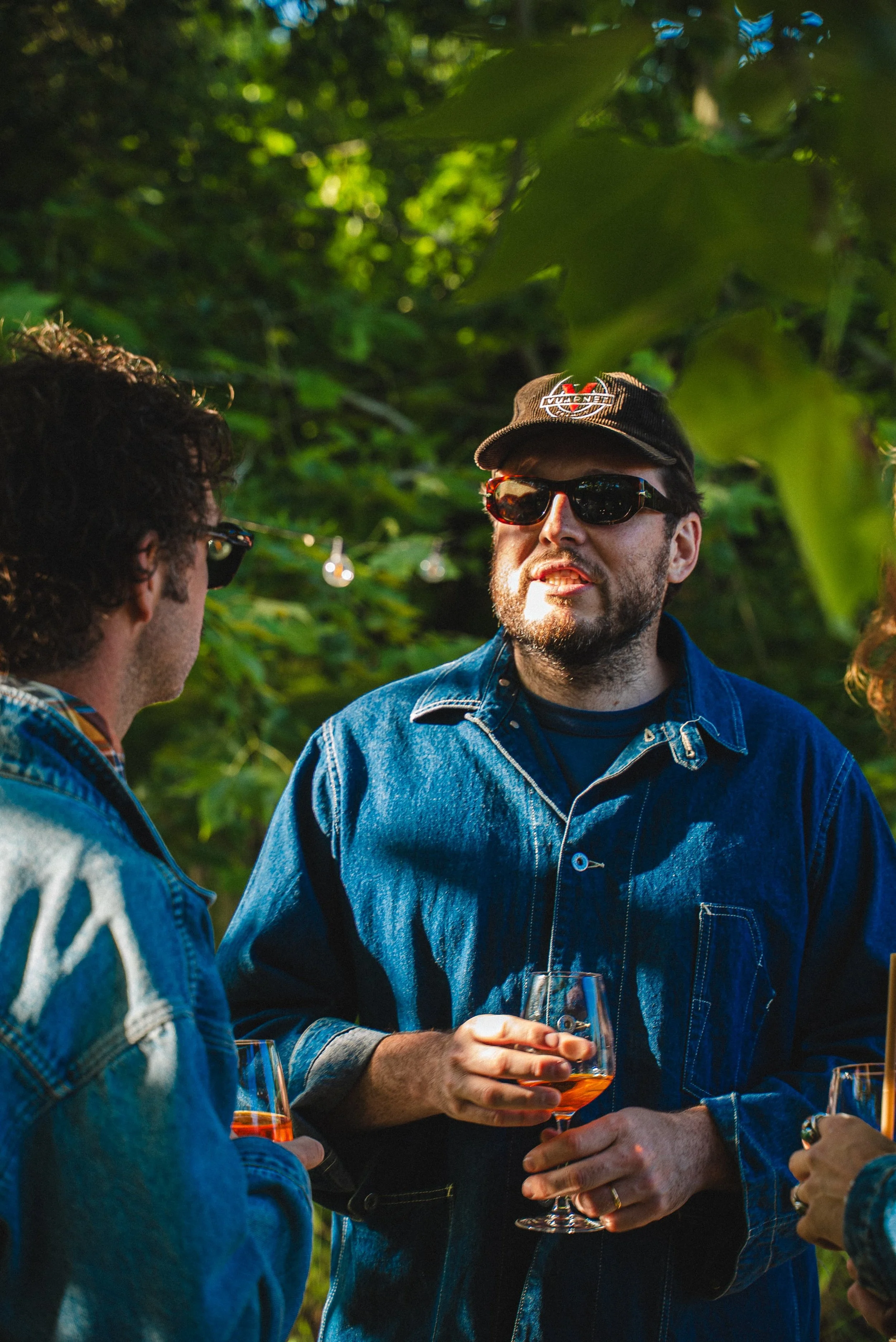 Groupe de personnes en conversation en extérieur, portant des vêtements en denim, avec un homme portant une casquette et des lunettes de soleil, tenant un verre de boisson colorée.