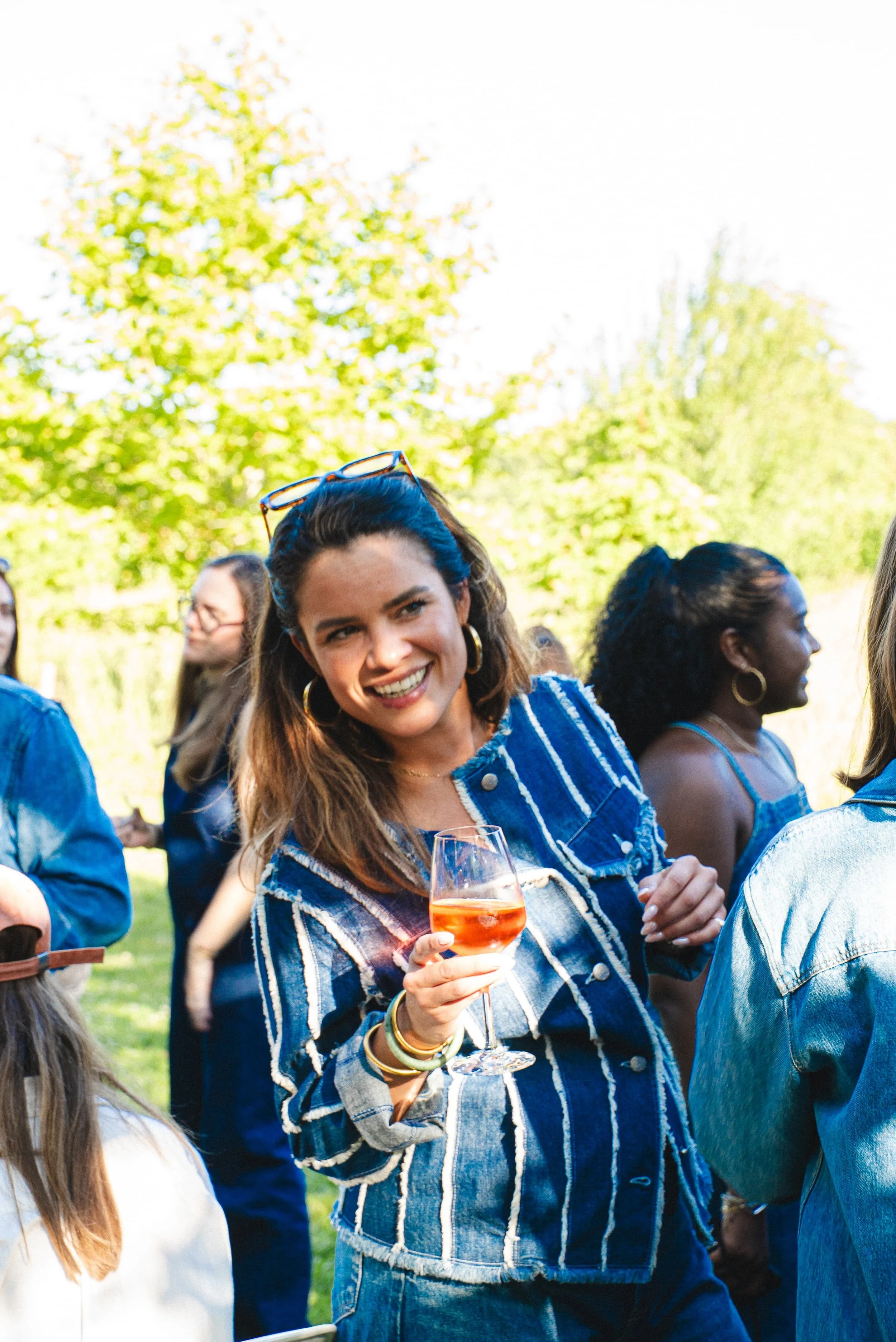 Femme souriante tenant un verre de vin rosé lors d'une réunion en plein air avec d'autres personnes.