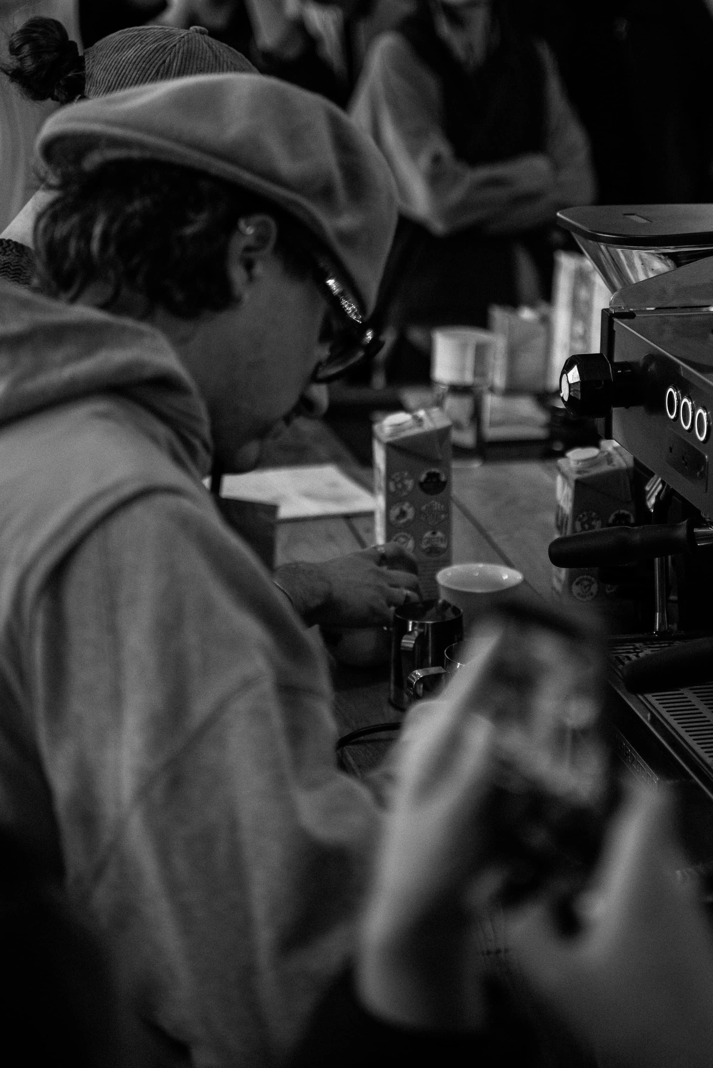 Une personne portant un bonnet et des lunettes, travaillant dans ce qui semble être un café ou une cuisine, entourée d'ustensiles et de machines à café.