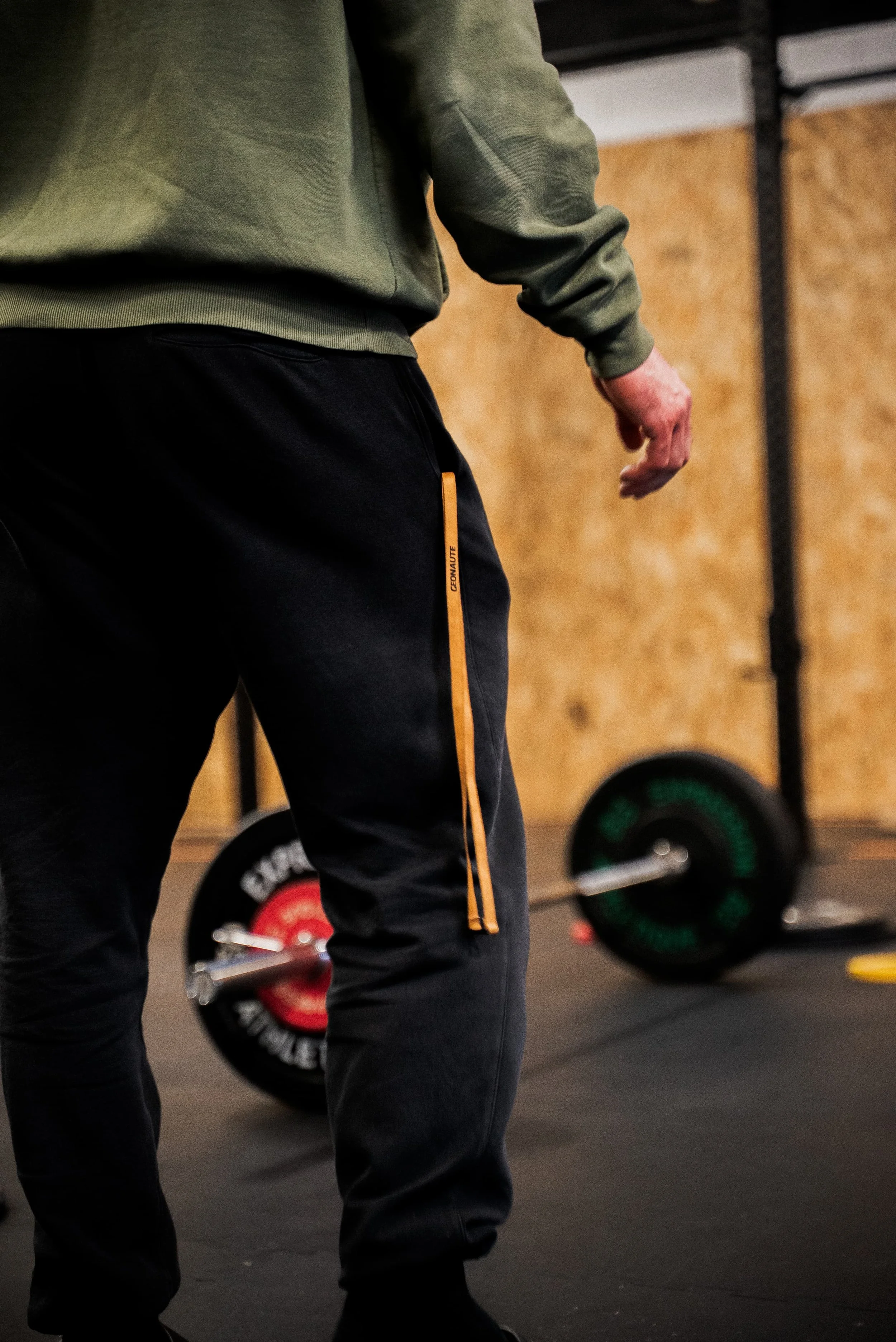 Vue rapprochée de la taille d'une personne portant un survêtement gris foncé et un pantalon noir dans une salle de sport. En arrière-plan, des haltères et des disques de poids sont visibles.